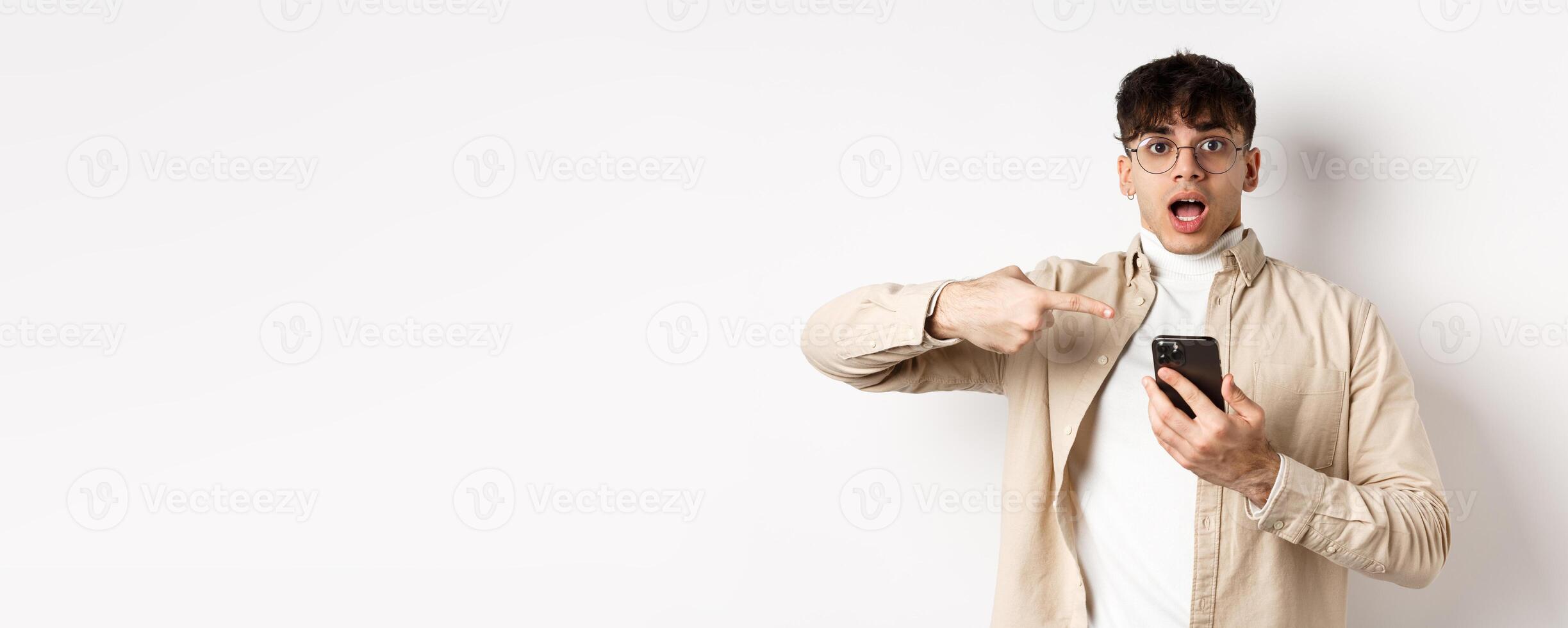 Real people. Amazed young guy checking out online offer on mobile phone, pointing at smartphone screen with excited face, standing on white background photo