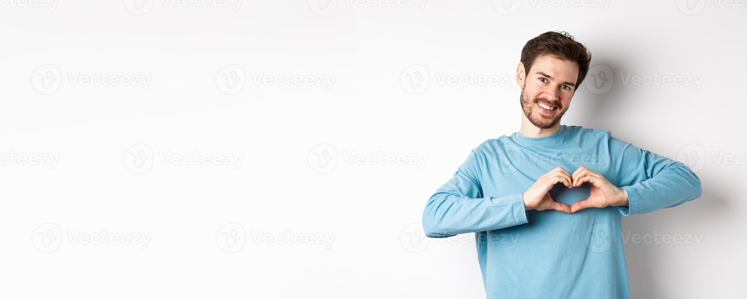 Handsome boyfriend saying I love you, showing heart gesture and smiling at camera, express love and romantic feeling, standing over white background photo