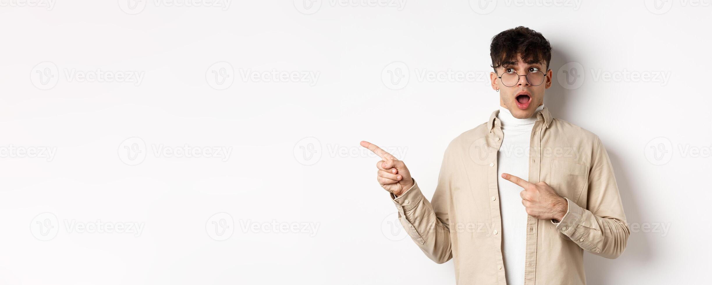 Amazed young man in glasses drop jaw, saying wow, pointing and looking left at awesome promo offer, stare with disbelief and impressed face, white background photo