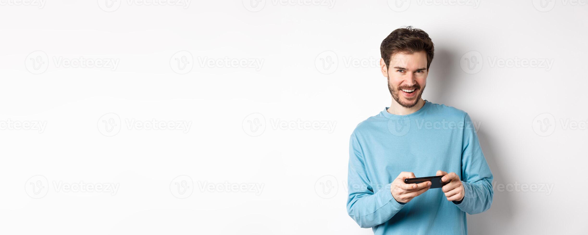 Image of handsome young man playing mobile game, holding smartphone horizontally and smiling at camera pleased, standing over white background photo