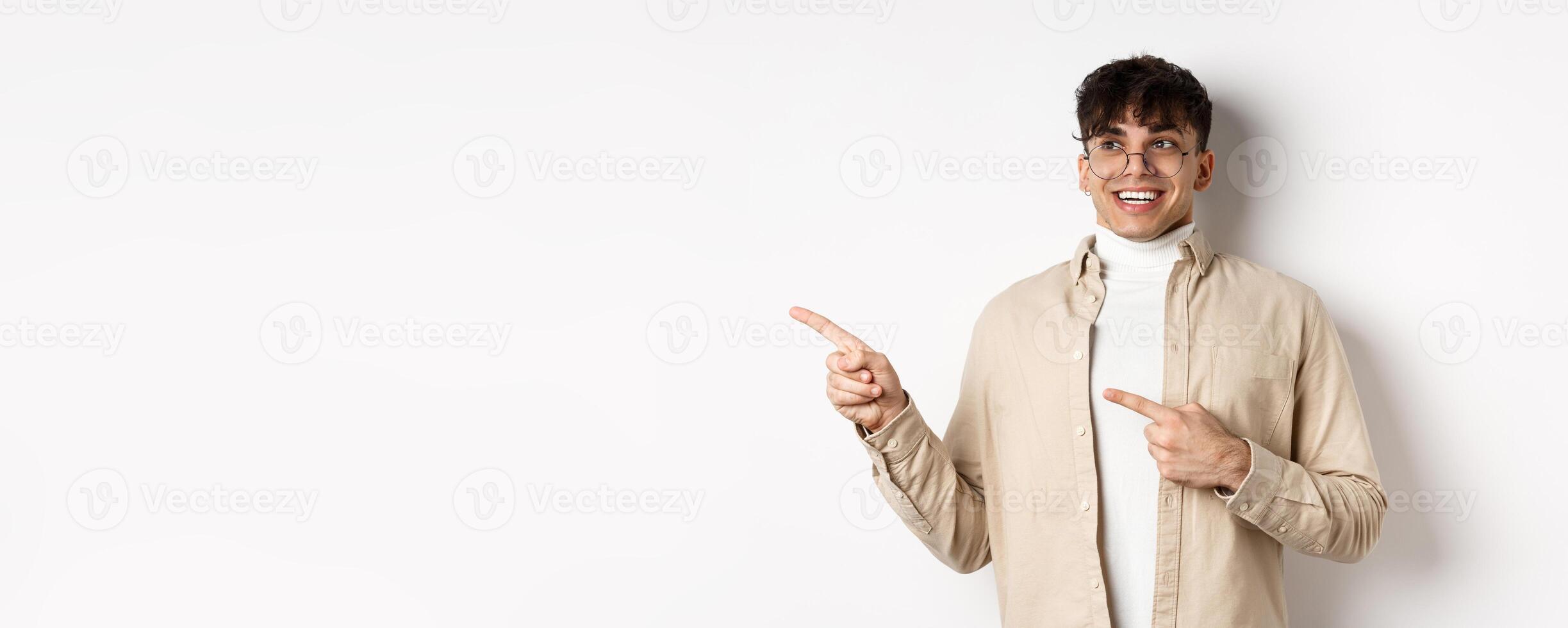 Excited and enthusiastic young man in glasses, pointing fingers and looking left with happy smile, standing in awe on white background photo