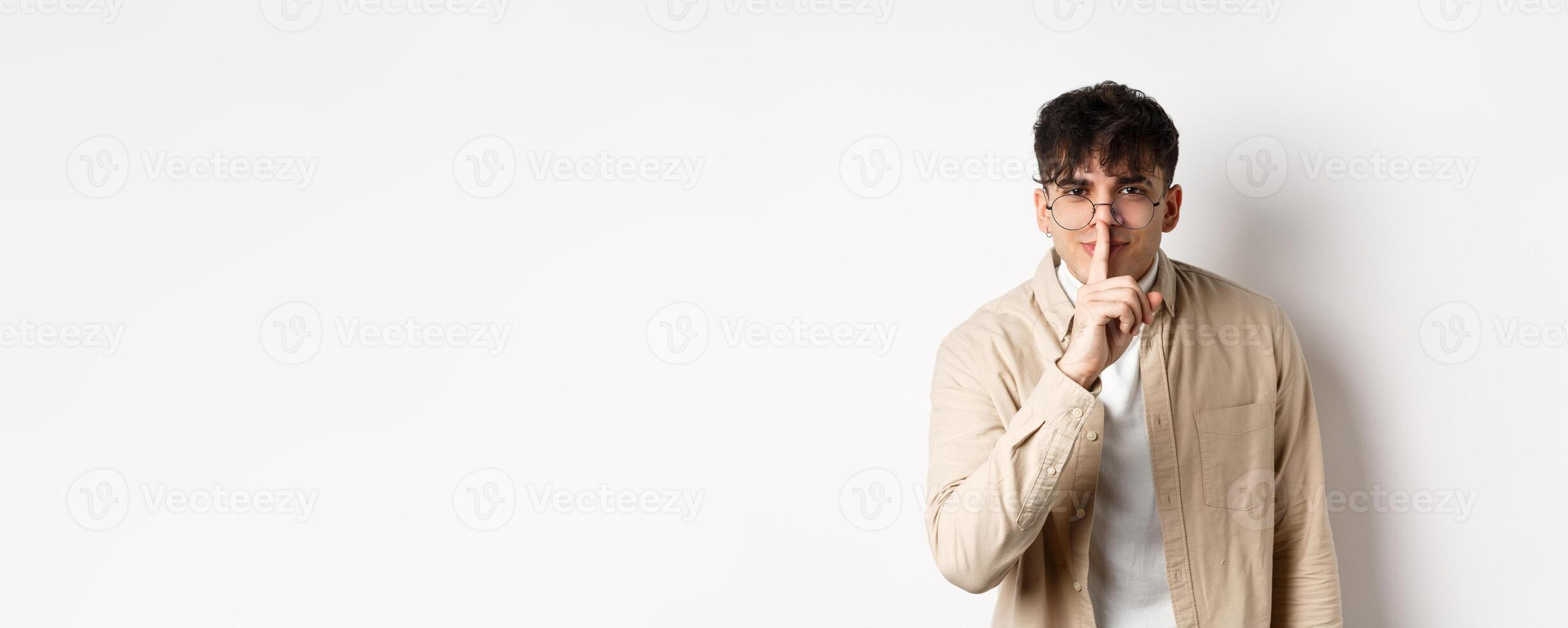 Cheerful handsome man in glasses share a secret, making surprise, lean at camera with hush gesture and gentle smile, telling to be quiet, show taboo sign, white background photo
