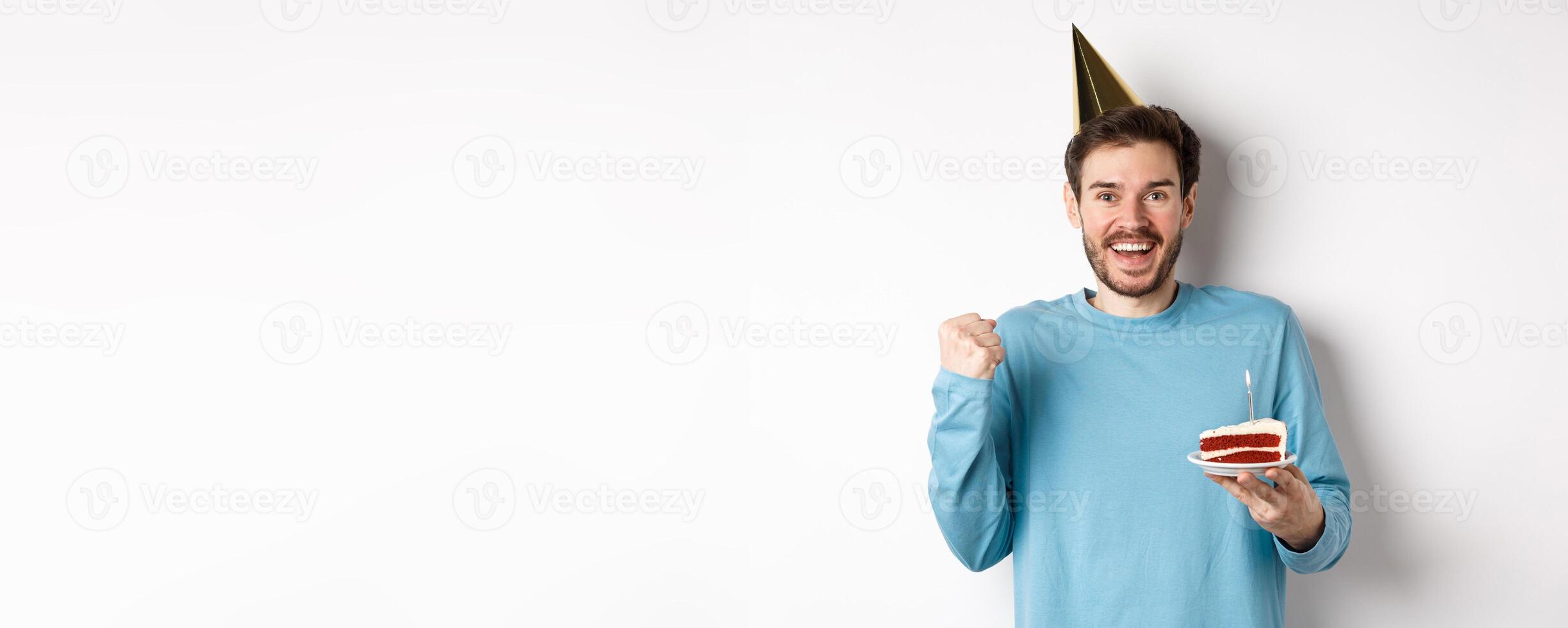 Celebration and holidays concept. Cheerful young man celebrating birthday in party hat, saying yes and fist pump in joy, holding bday cake, white background photo