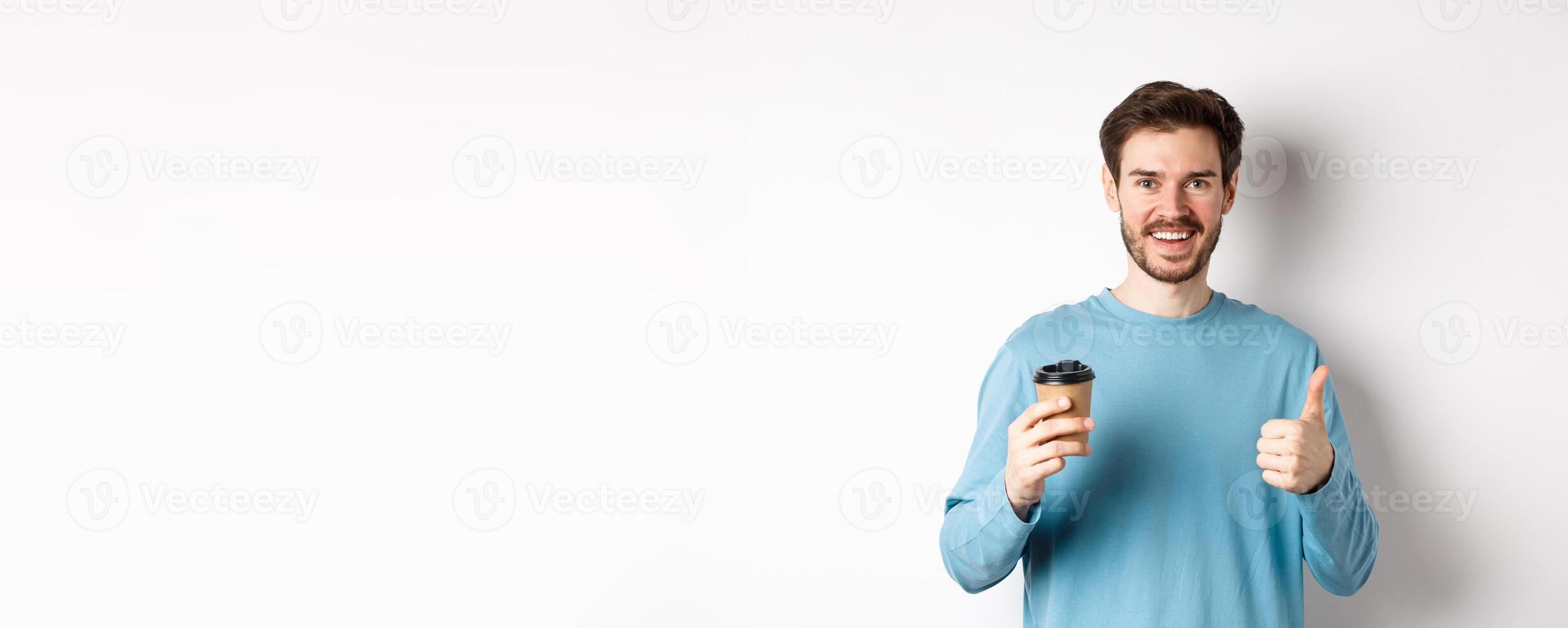 Cheerful young man drinking good coffee, holding paper cup and showing thumb up, recommending cafe shop, standing over white background photo