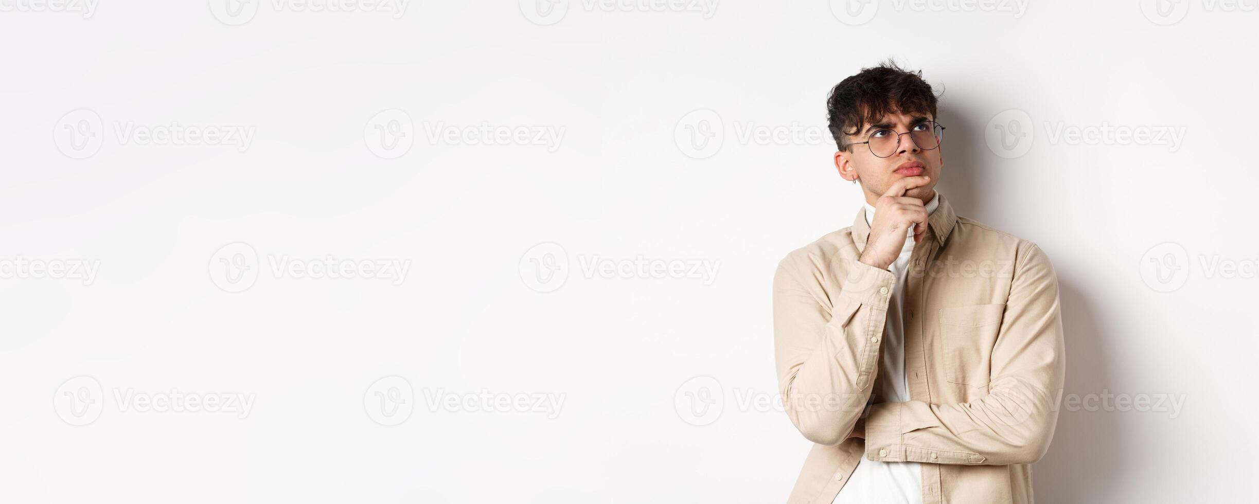 Handsome hipster guy in glasses look pensive at empty space, touching chin and frowning, stare up thoughtful, making choice with doubtful face, standing on white background photo