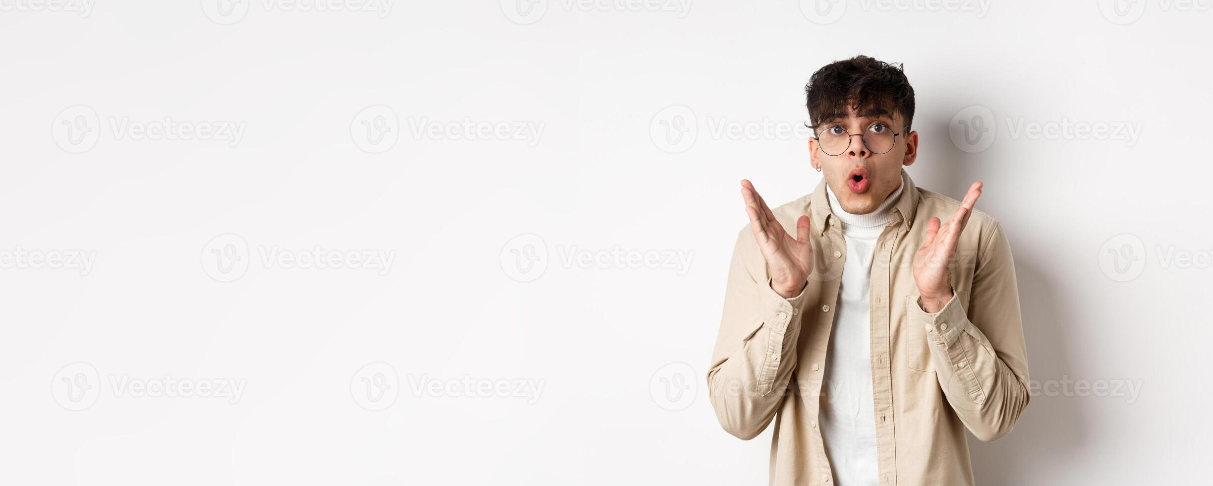 Portrait of surprised hipster guy in glasses checking out awesome promo, say wow and gasping amazed, raising hands up with disbelief, standing on white background photo