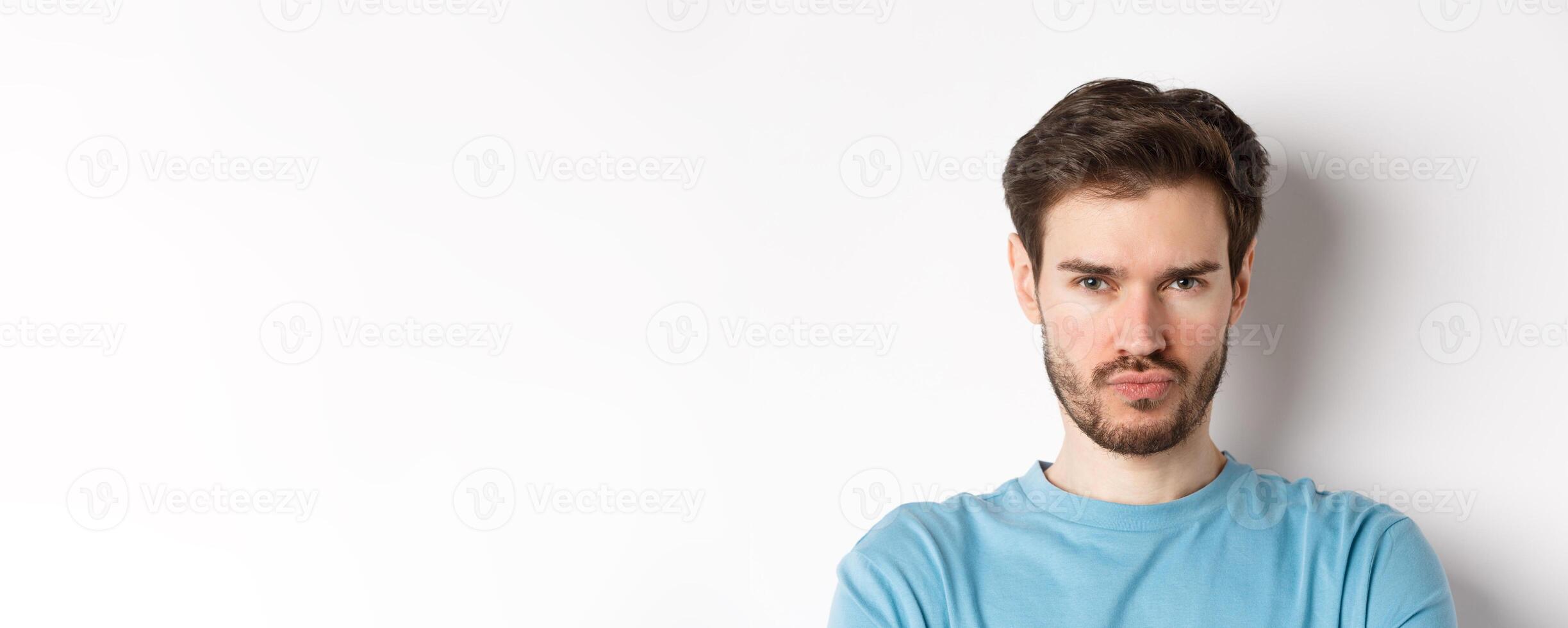 Close-up of grumpy young man grimacing, sulking and making offended face, standing upset over white background photo