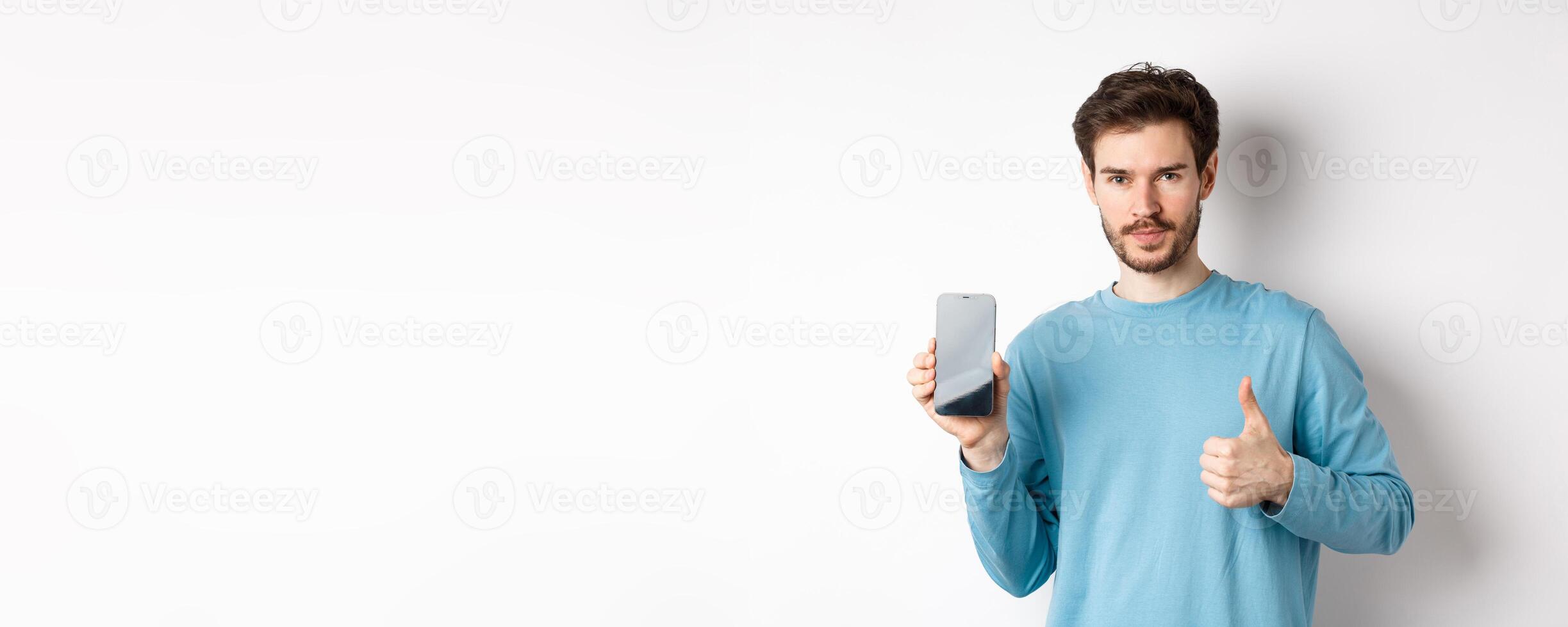 Young man showing empty smartphone screen and thumb up, recommend mobile application or online shopping offer, standing over white background photo