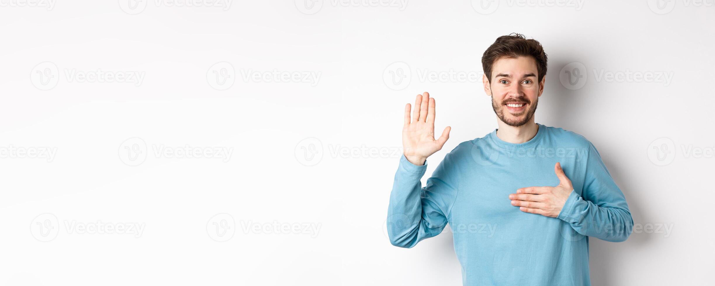Smiling hosent man raising arm and hold hand on heart, making promise tell truth, swear or give oath, standing over white background photo