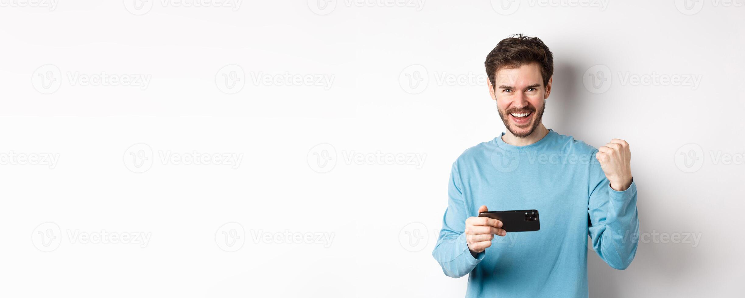 Lucky man winning in mobile game, holding smartphone and saying yes, triumphing with fist pump, standing over white background photo