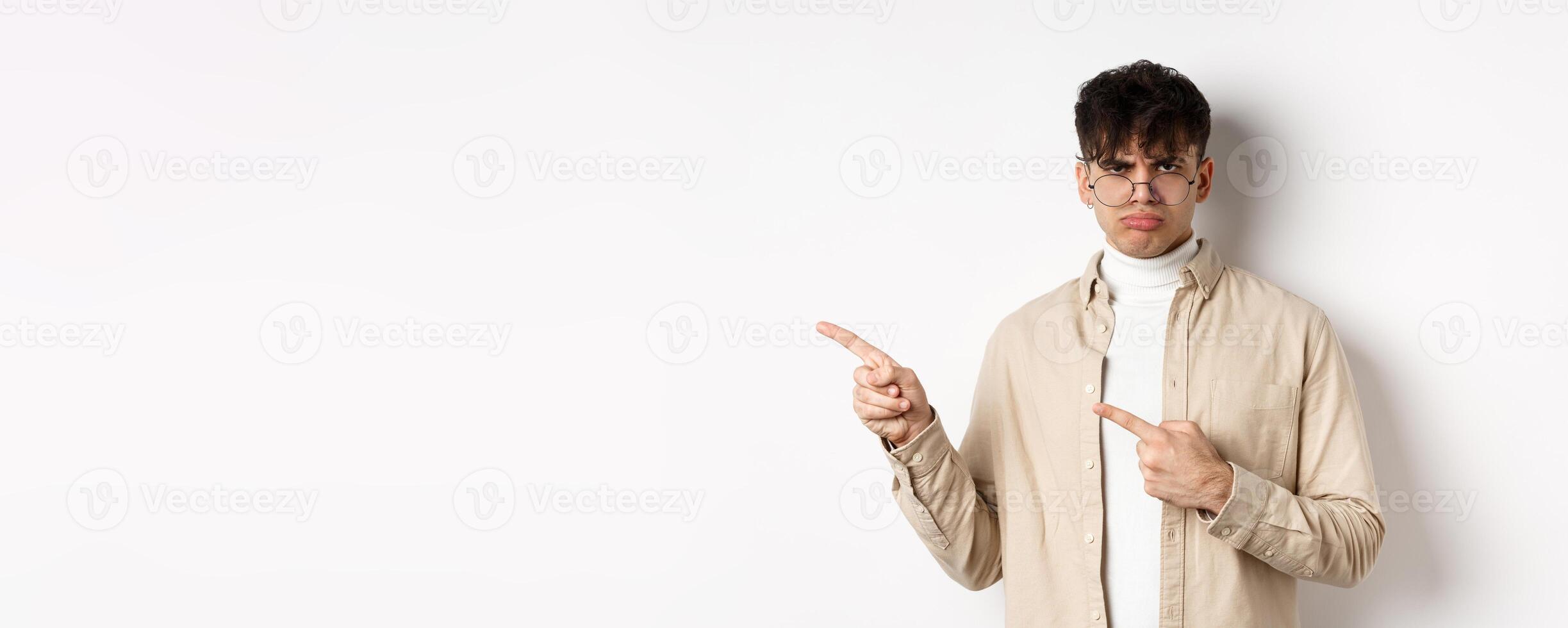 Sad and grumpy boy pointing fingers left, sulking with childish expression, complaining on something bad, standing on white background photo