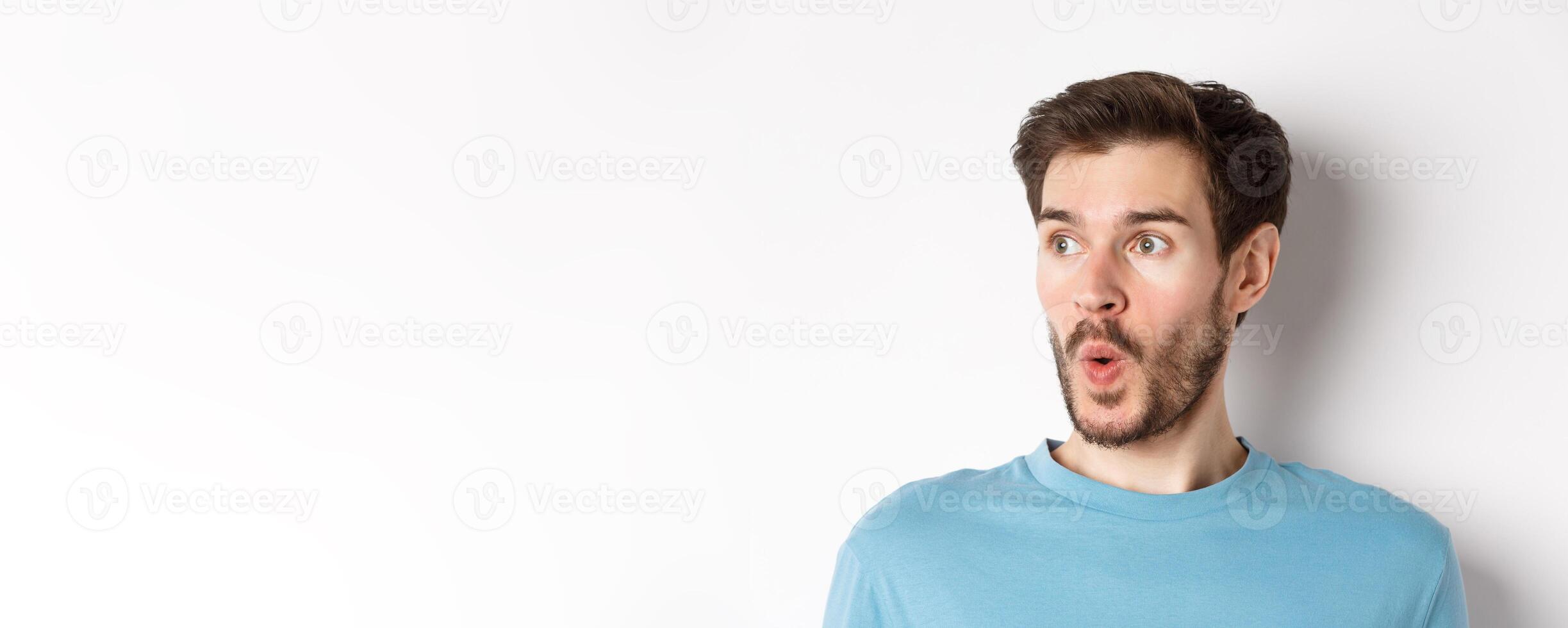 Close-up of impressed caucasian man saying wow, looking left amazed, checking out promotion deal, white background photo