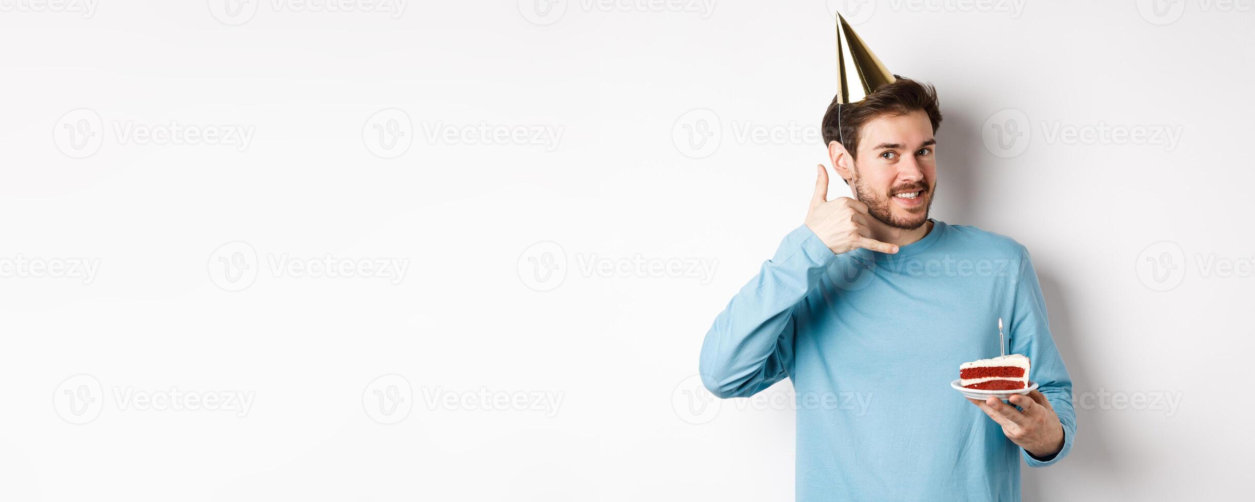 Celebration and holidays concept. Cheerful smiling man in party hat, celebrating birthday with bday cake, showing call me phone gesture near ear, white background photo