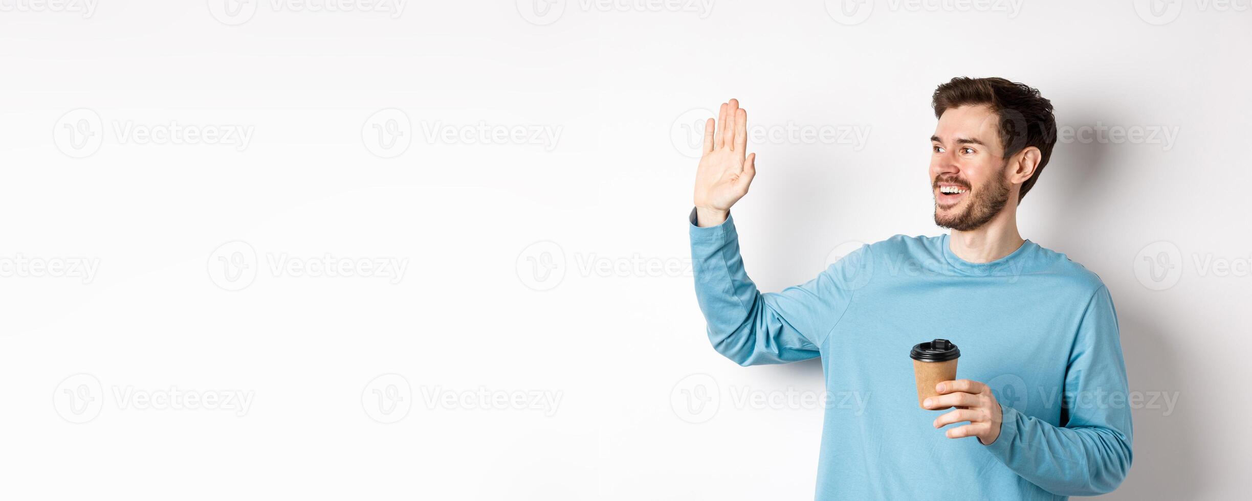 Friendly young man drinking coffee from takeaway cup and saying hello, waving hand left and smiling, standing over white background photo