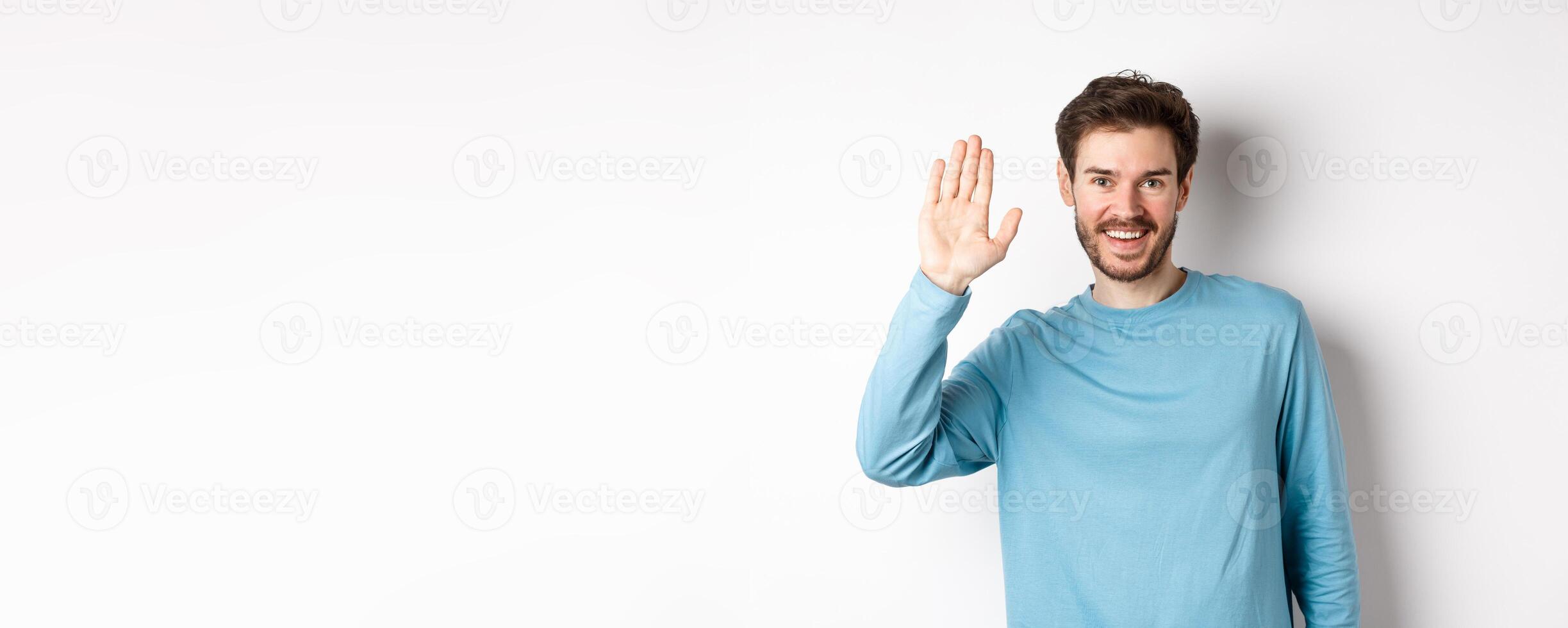 Cheerful young man with beard saying hello, looking friendly and waving hand to greet you, standing over white background photo