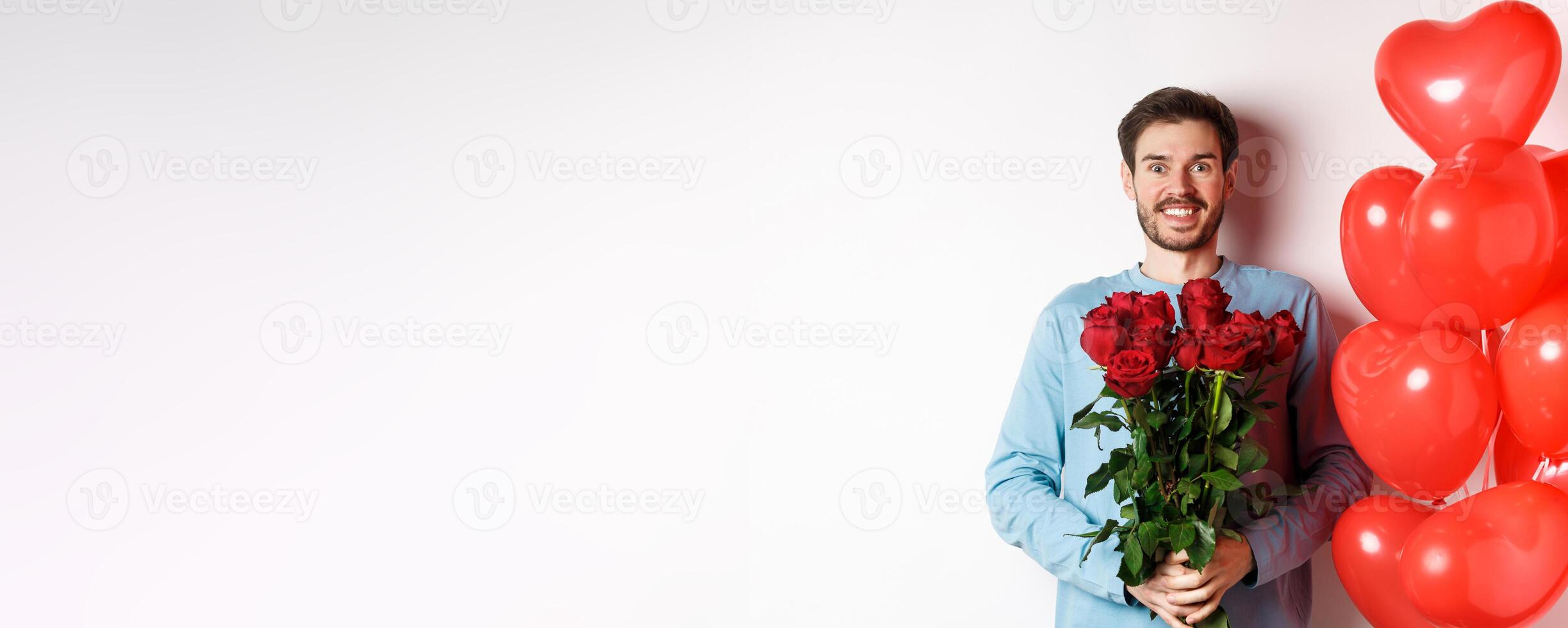 Valentines day romance. Excited young man with bouquet of red roses and heart balloons smiling at camera, bring presents for lover on valentine date, standing over white background photo
