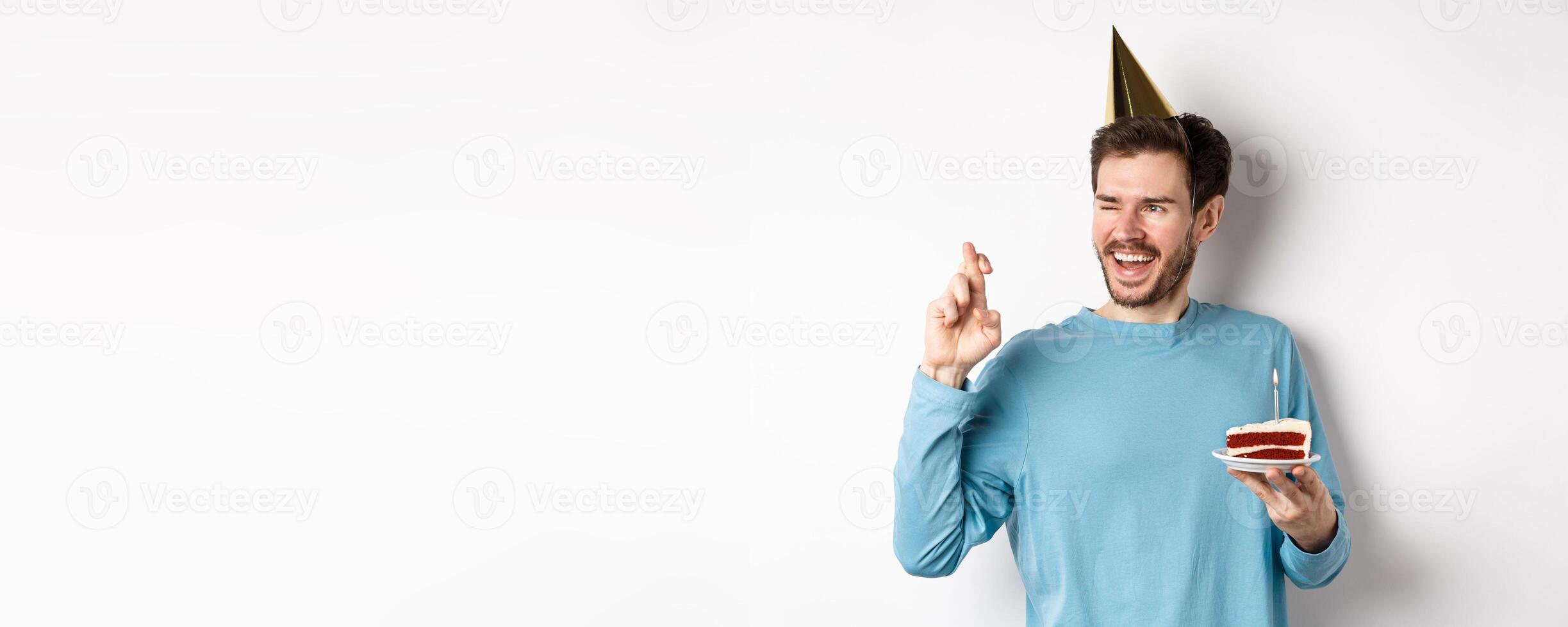 Celebration and holidays concept. Cheerful young man making wish on birthday, holding fingers crossed and wearing party hat, standing with bday cake, white background photo