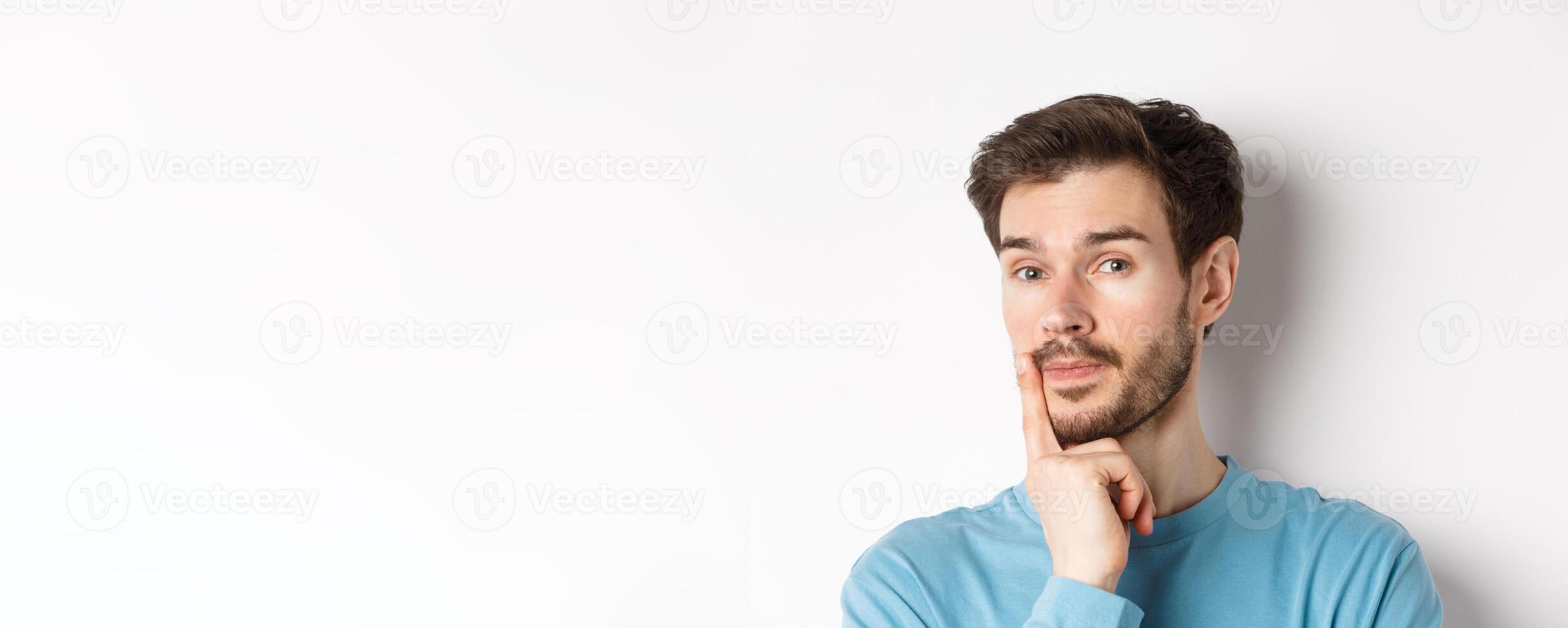 Image of thoughtful young man with beard, making choice, touching lip and looking pensive at camera, deciding something, white background photo