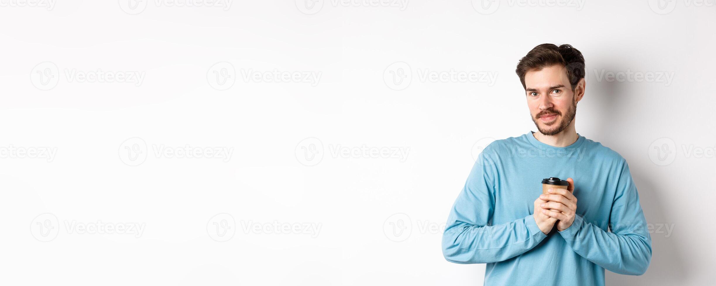 Handsome caucasian man warm-up hands with cup of coffee, smiling at camera and looking tenderly, standing on white background photo