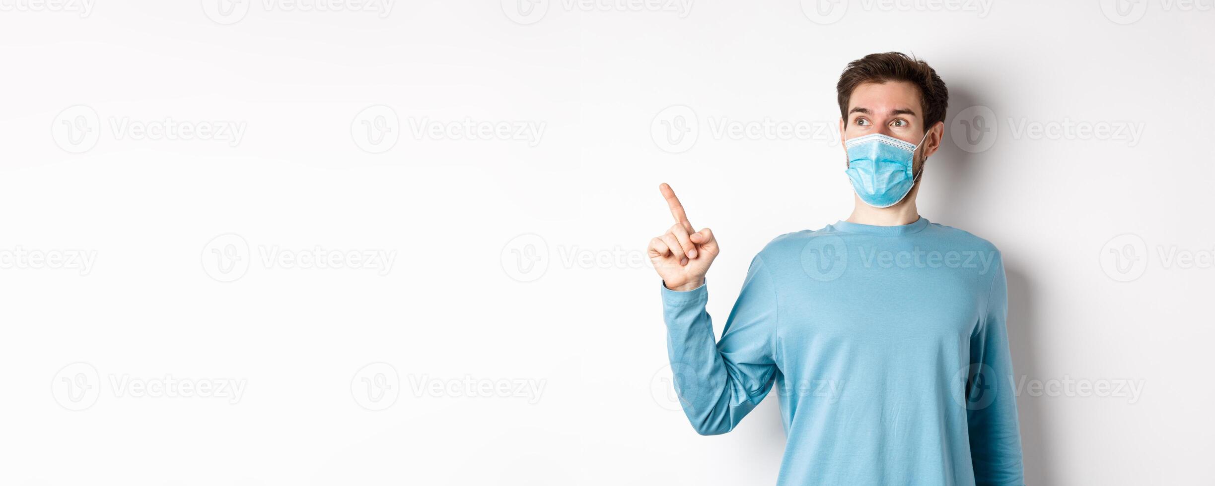 Coronavirus, health and quarantine concept. Surprised guy looking in awe and pointing at upper left corner, standing in medical mask and sweatshirt over white background photo