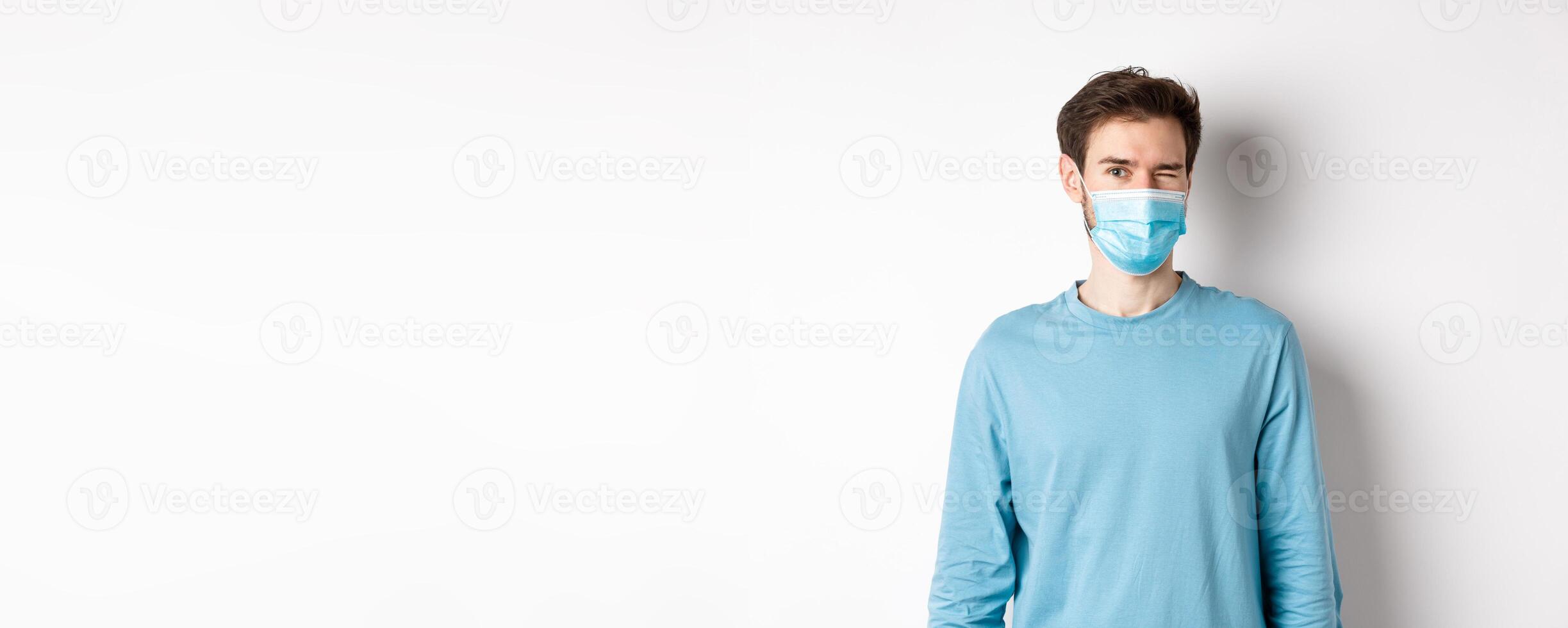Covid-19, pandemic and social distancing concept. Cheerful caucasian man in face mask winking at camera, looking happy, standing over white background photo