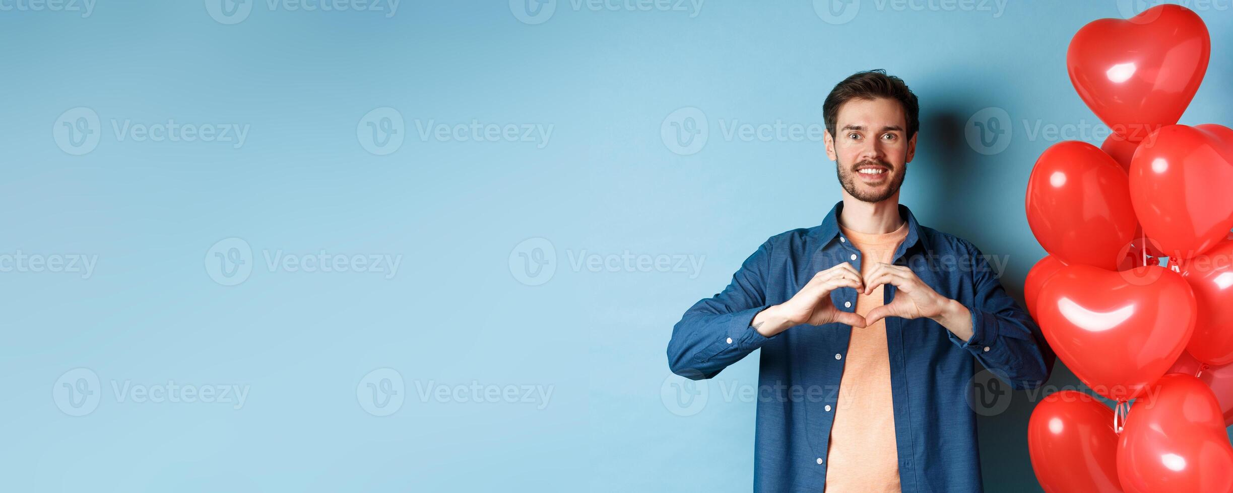 Happy valentines day. Boyfriend in love showing heart gesture to lover and smiling, standing near red balloons on blue background photo