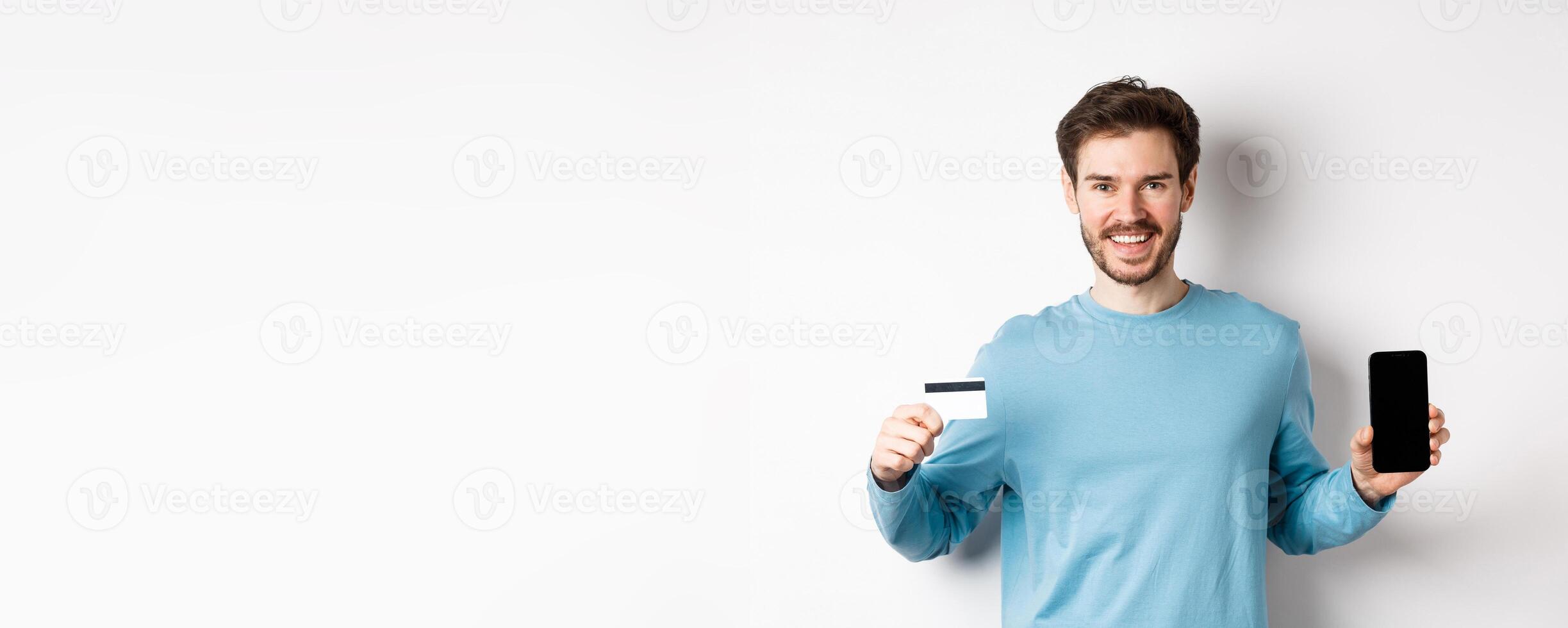 Smiling caucasian man showing plastic credit card with mobile phone screen. Guy recommending online banking app, standing on white background photo