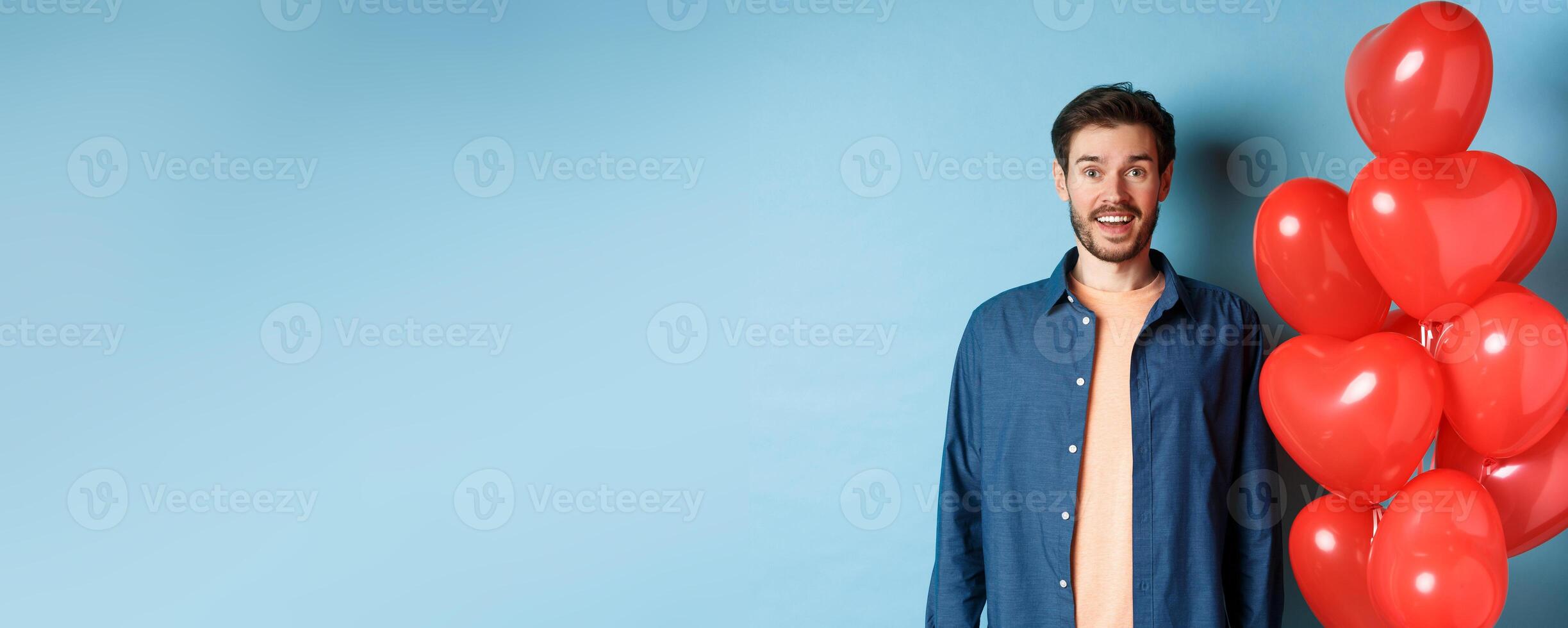 Happy valentines day. Excited smiling guy standing near red hearts balloons and looking at camera, blue background photo