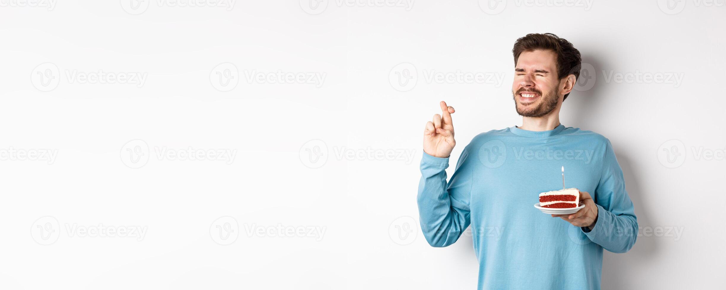 Celebration and holidays concept. Young man celebrating birthday, cross fingers for good luck, making wish on bday cake with lit candle, standing over white background photo