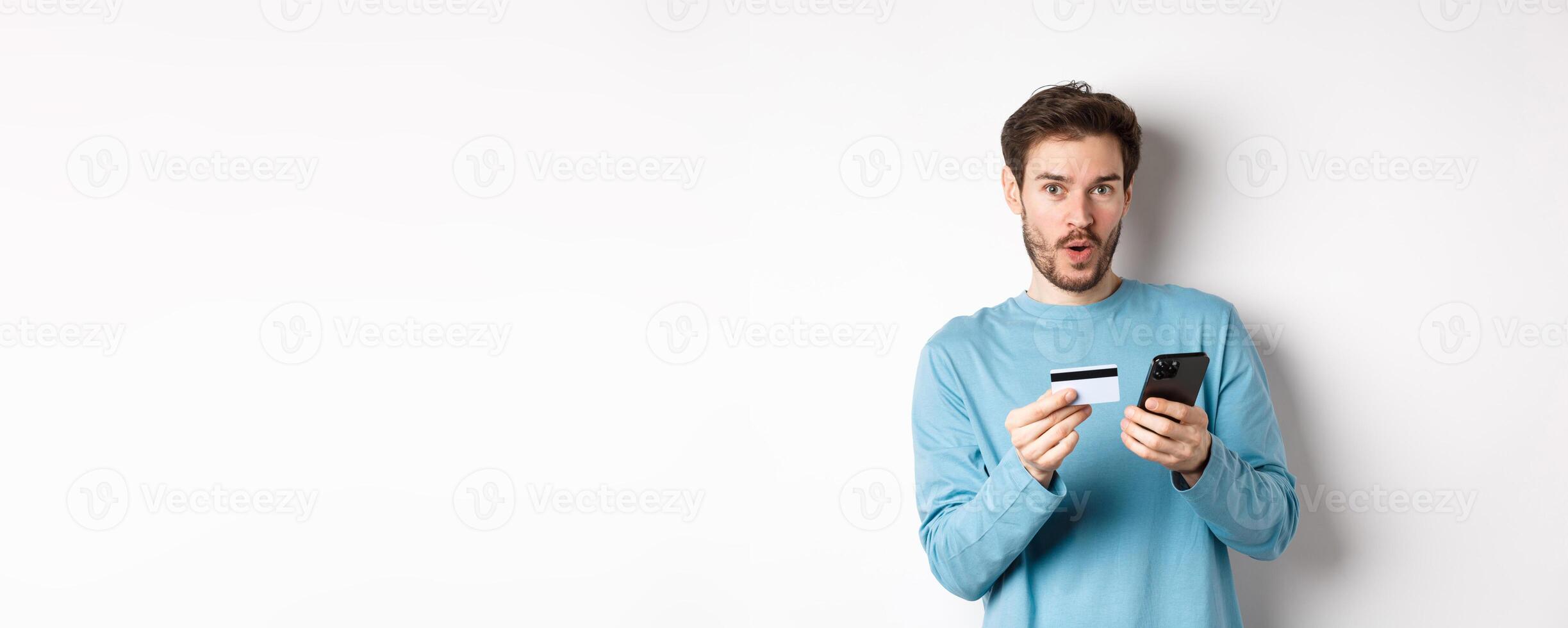 E-commerce and shopping concept. Excited man looking amused while making order online, holding plastic credit card and smartphone, standing on white background photo