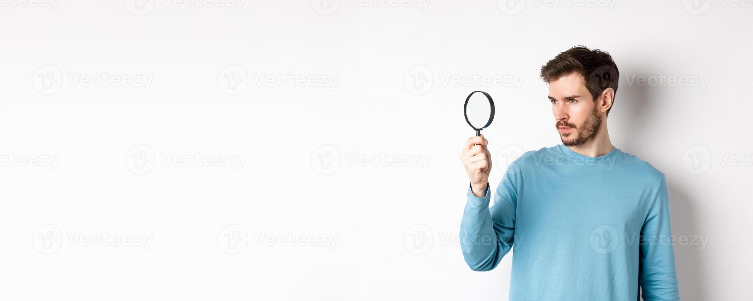 Young handsome man look through magnifying glass with curious face, investigating or searching for something, standing on white background photo