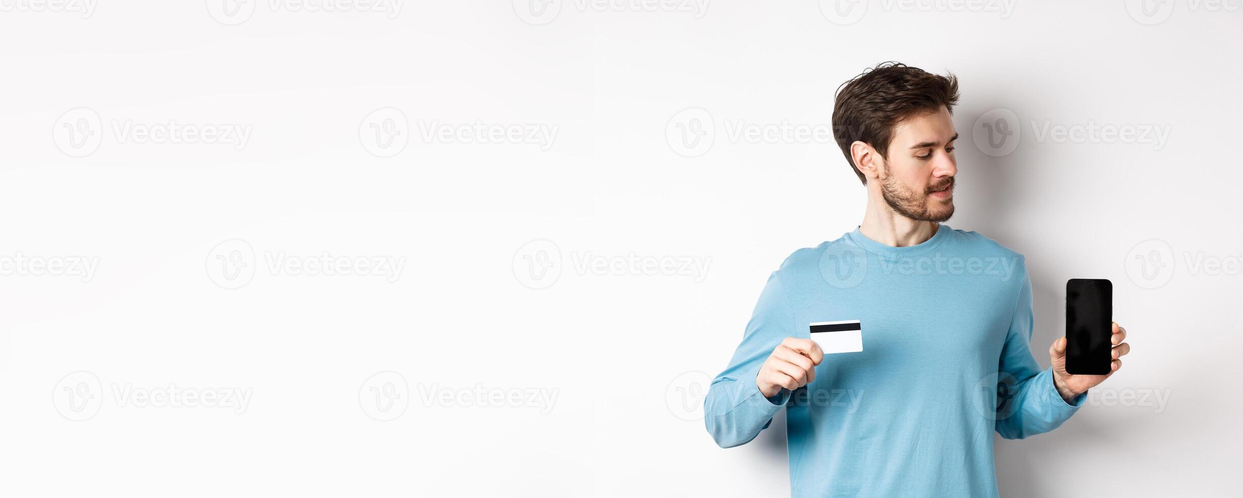 Mobile banking. Handsome caucasian man showing plastic credit card and looking at blank mobile screen, standing over white background photo