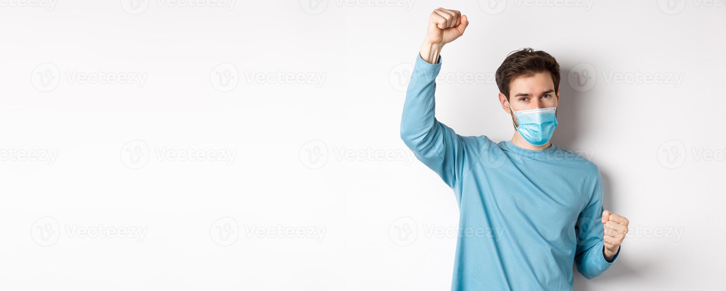 Covid-19, pandemic and social distancing concept. Happy and lucky guy in medical mask winning, raising hands up and celebrating achievement, white background photo