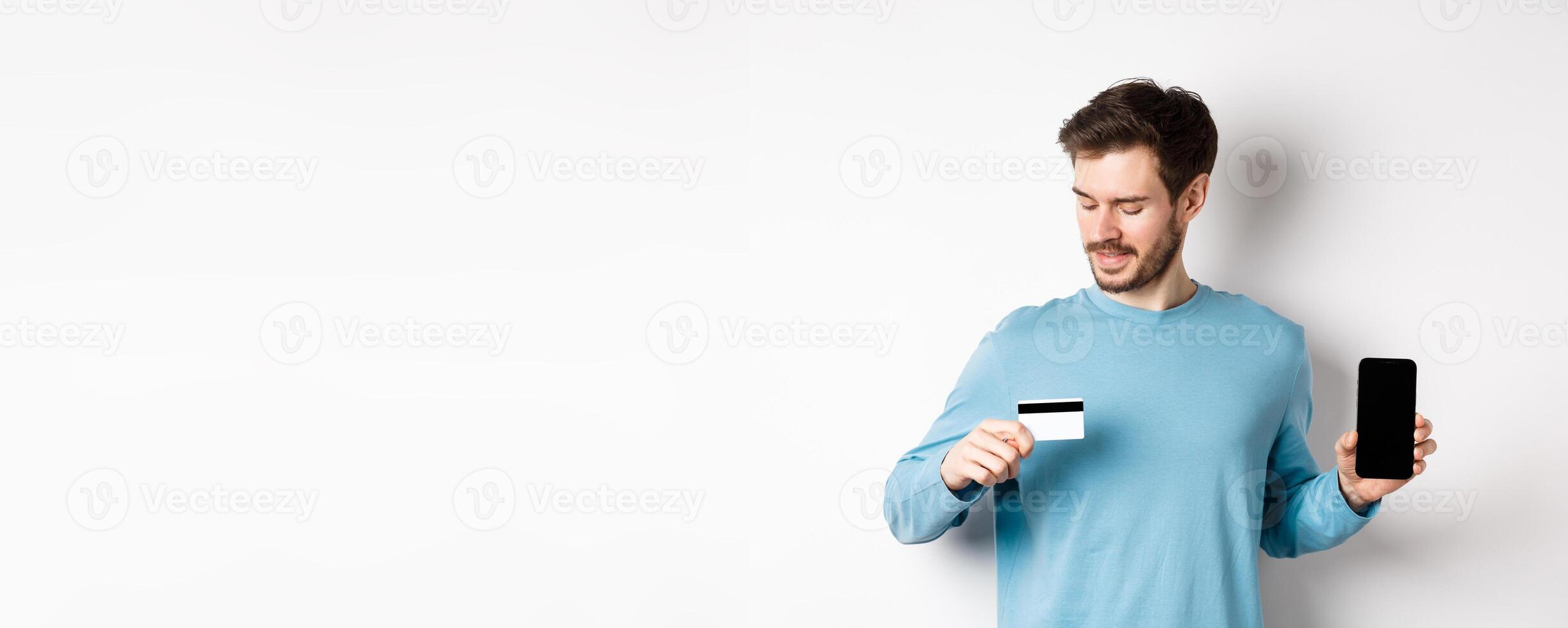 Handsome young man with beard, showing empty smartphone screen and looking at plastic credit card, standing over white background photo