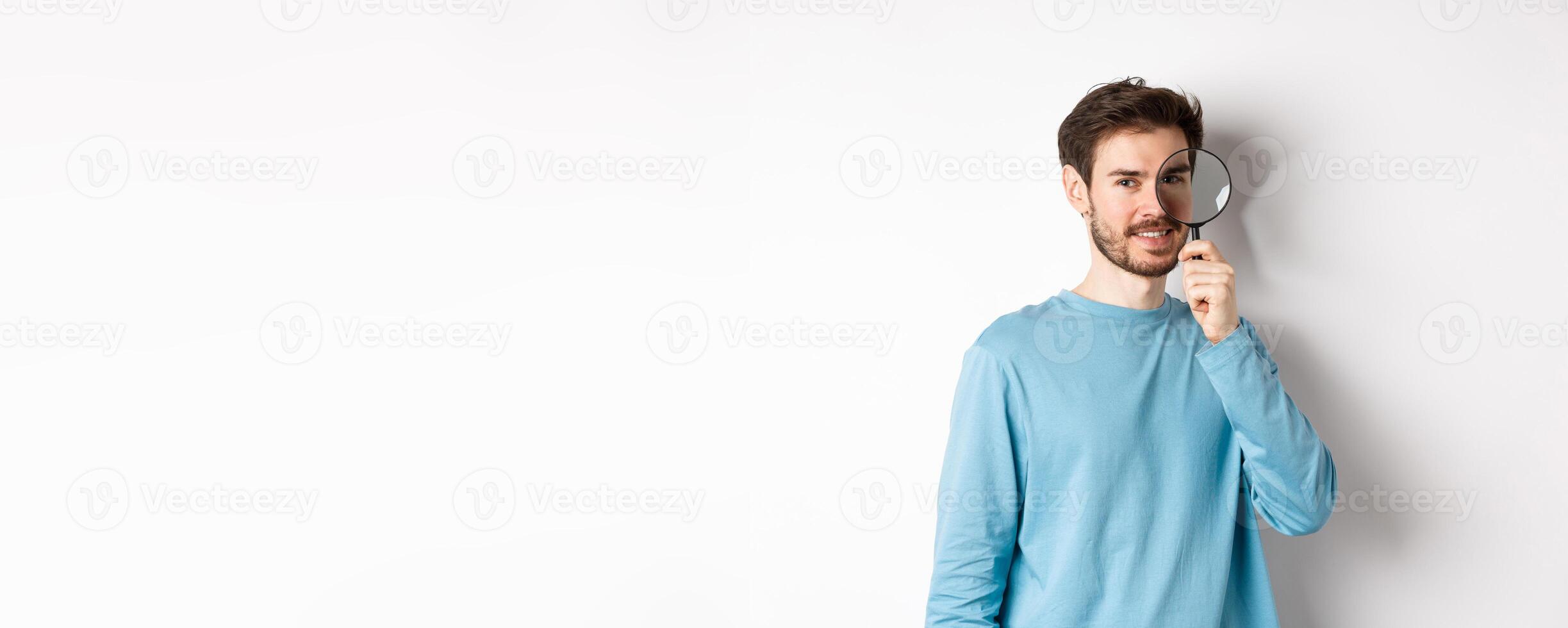 Smiling young man detective looking through magnifying glass, searching for promo offer, standing on white background photo