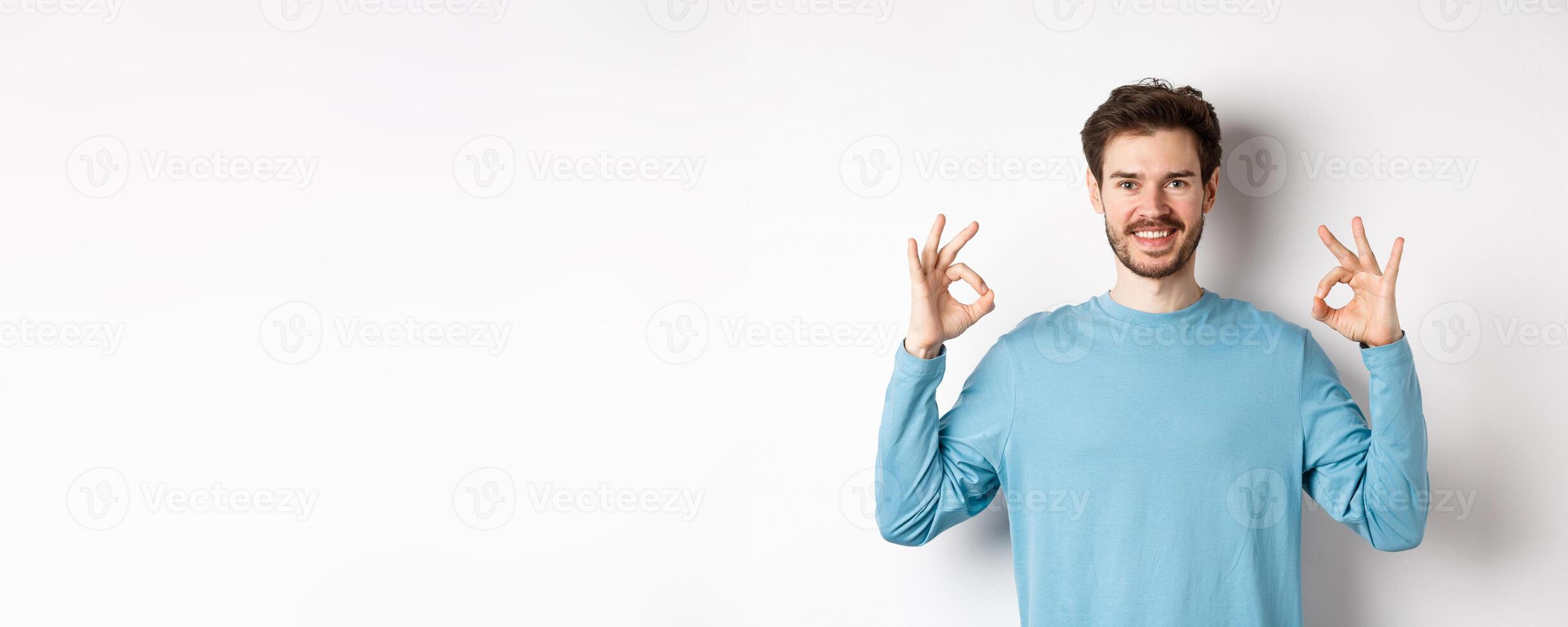 Very good. Smiling handsome guy showing okay signs and looking satisfied, standing over white background, approve excellent choice photo