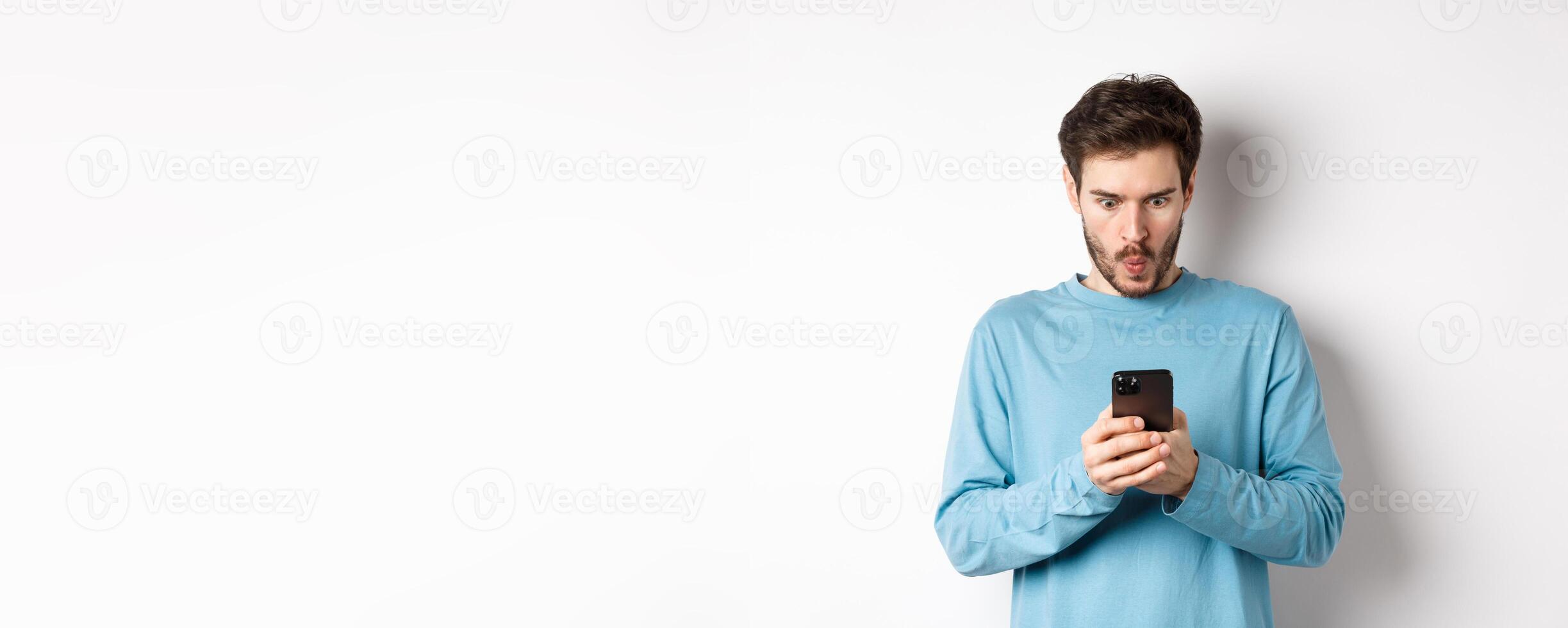Amazed young man staring at smartphone screen and saying wow, looking at online offer, standing on white background photo