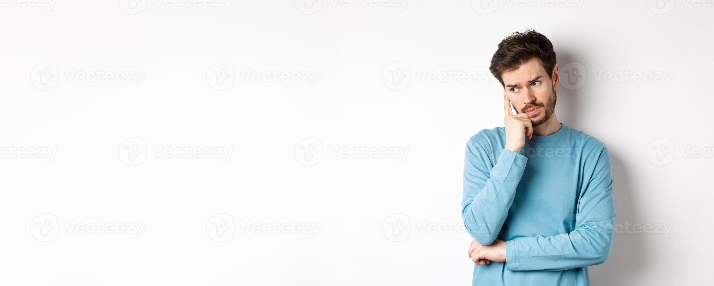 Worried young man with beard in sweatshirt, looking away pensive and thinking, standing troubled against white background photo