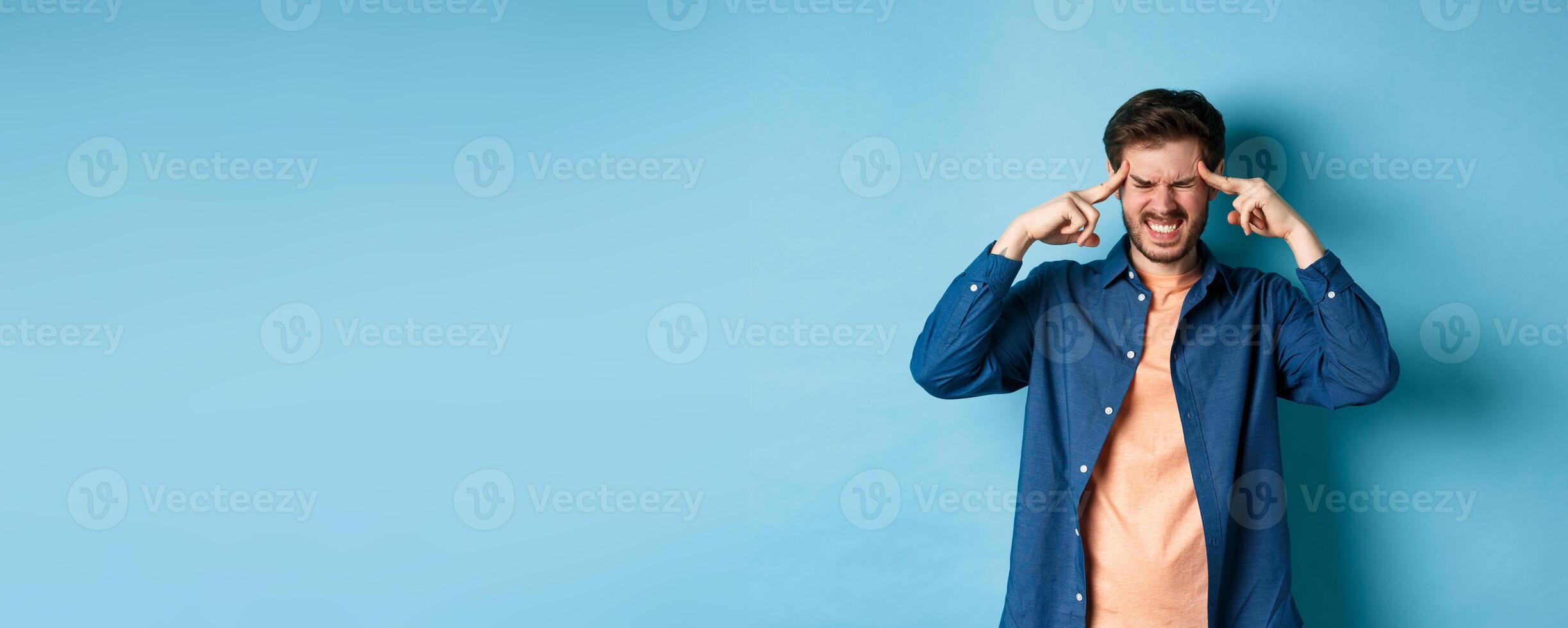 Man feeling sick, touching head and grimacing from painful migraine, having headache or feel dizzy, standing on blue background photo
