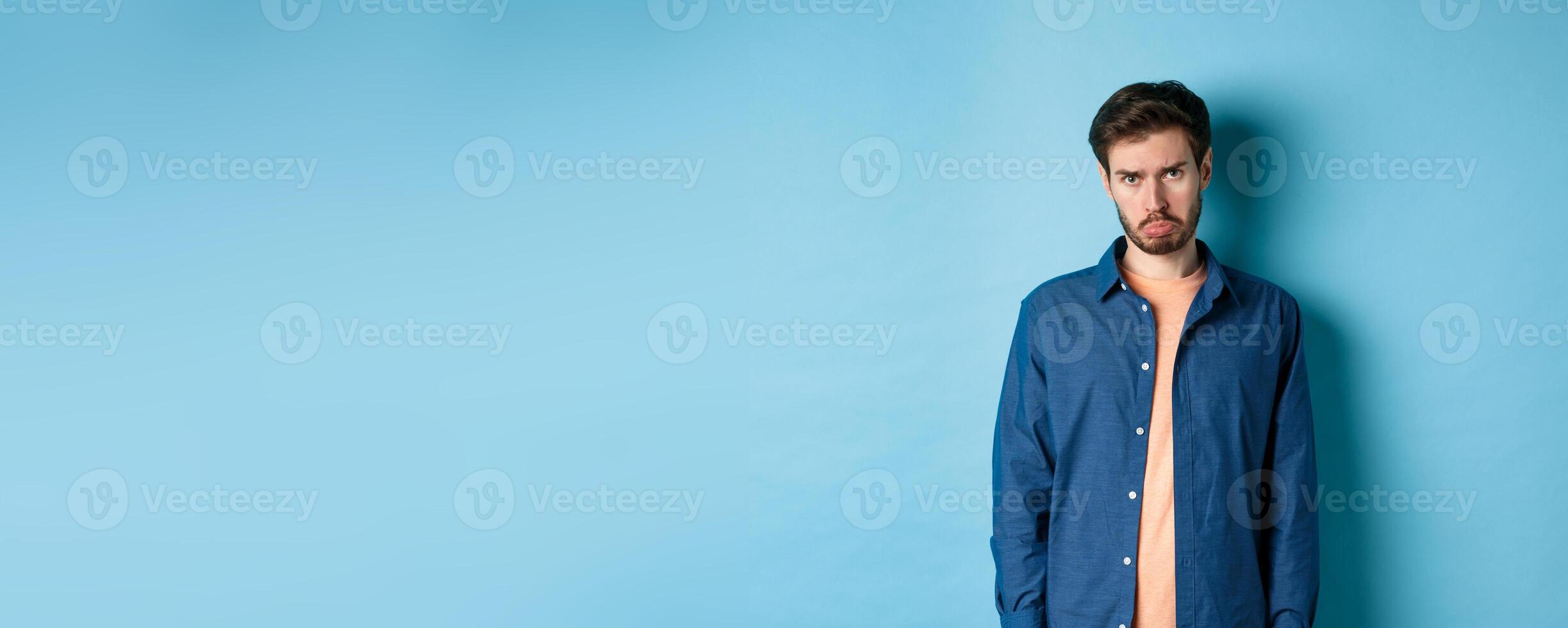 Sad and gloomy young man sulking, pulling miserable face and looking pleading at camera, standing on blue background photo