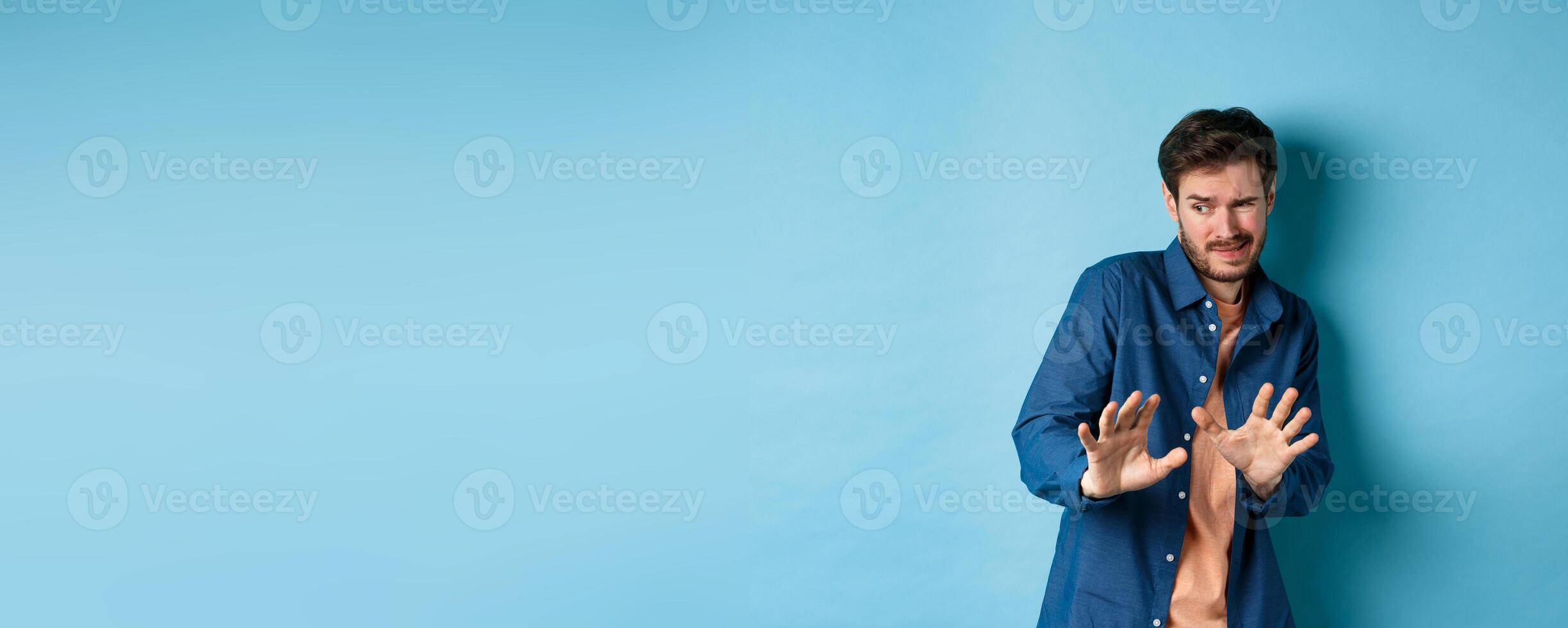 Man cringe and stay away from something nasty, looking aside and stretch out hands to reject bad deal, standing on blue background photo