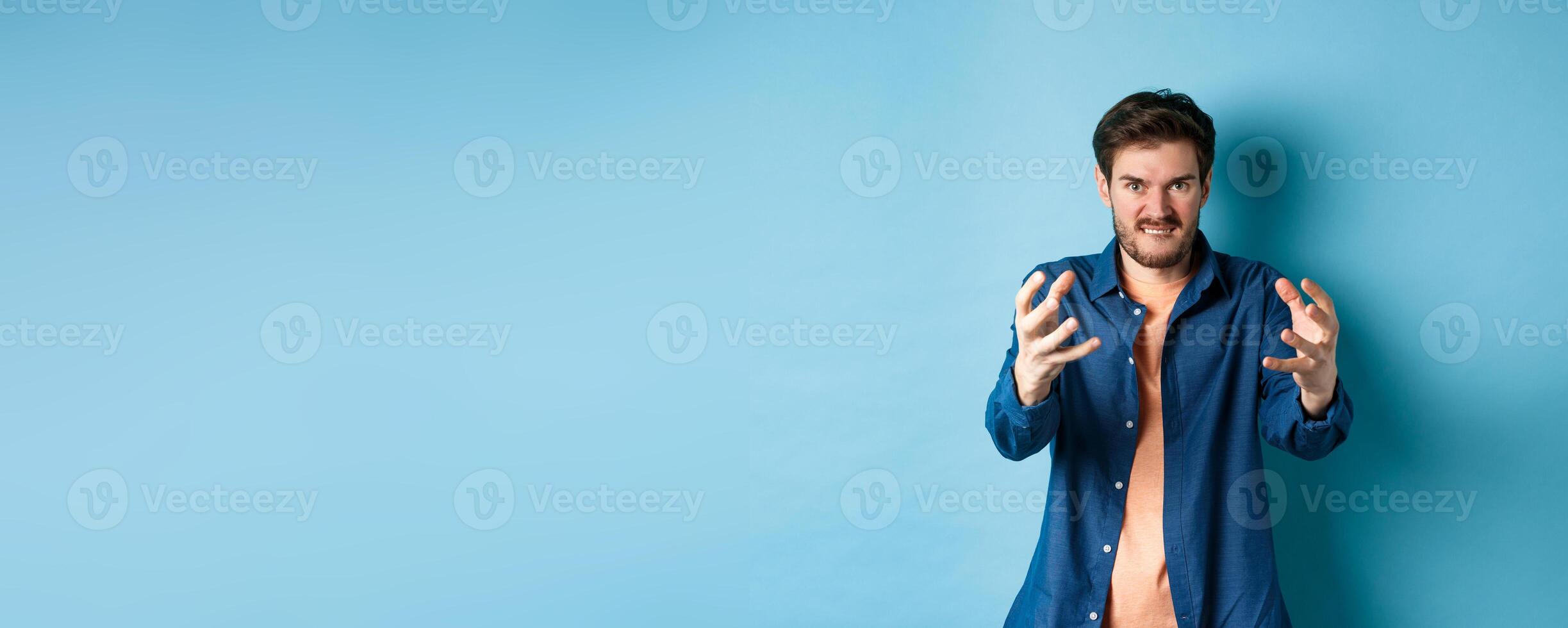 Angry tensed man wants to strangle someone, stretch out hands to choke enemy, biting lip and looking furious, standing on blue background photo