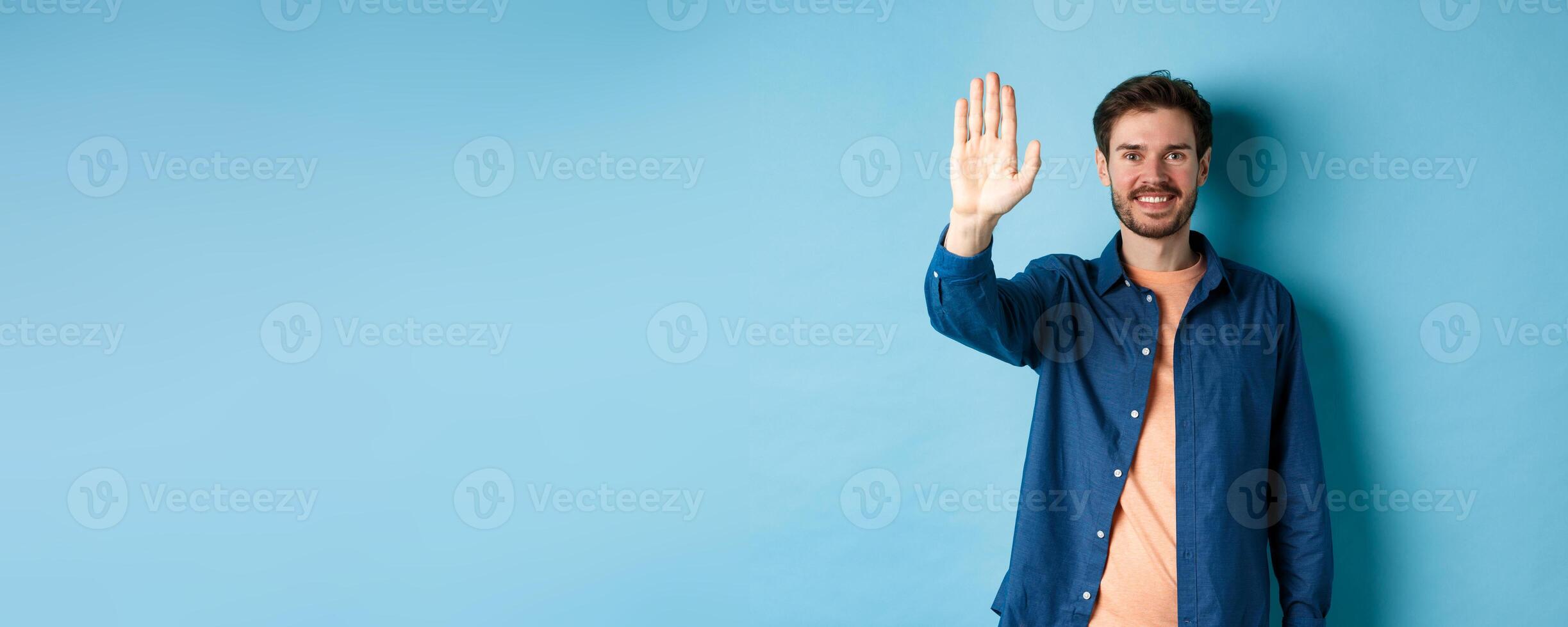 Friendly young man smiling and raising hand up for high five, waiving to greet or say hello, standing on blue background photo