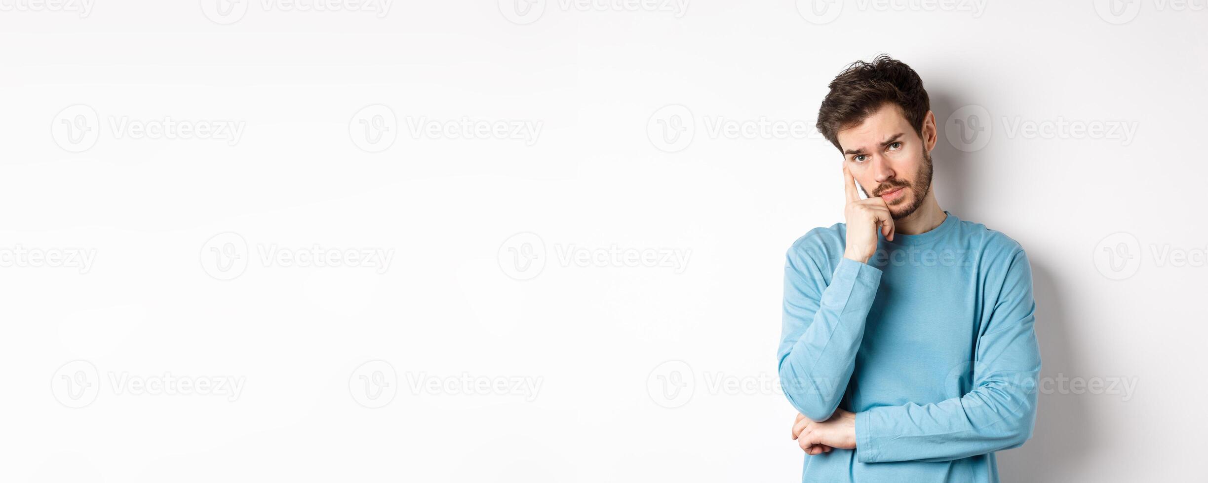 Skeptical young man listening to you with bored face, looking at camera reluctant, standing in sweatshirt over white background photo