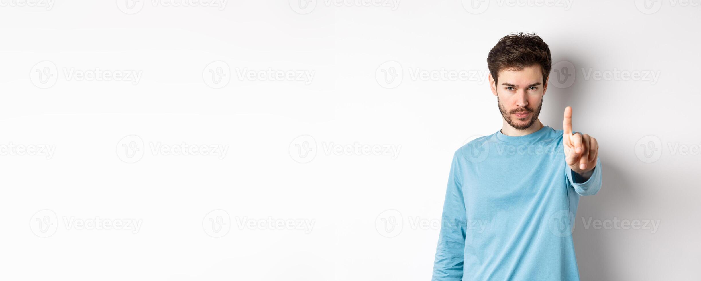 Image of serious young man with beard, shaking finger in disapproval, prohibit or forbid something, standing over white background and saying no photo