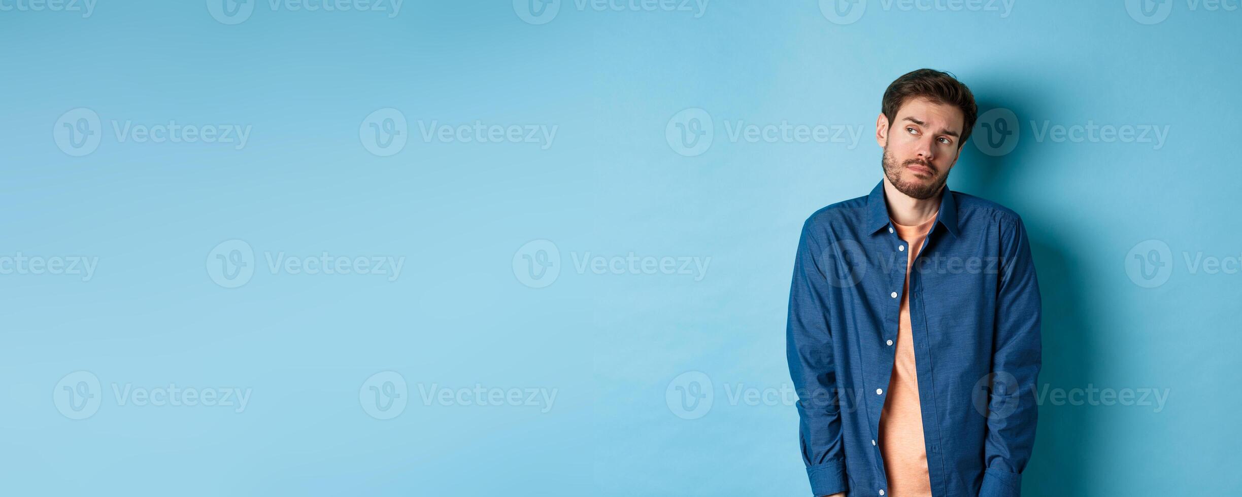 Reluctant and sad man feeling upset, looking aside at empty space with unamused face, standing on blue background photo