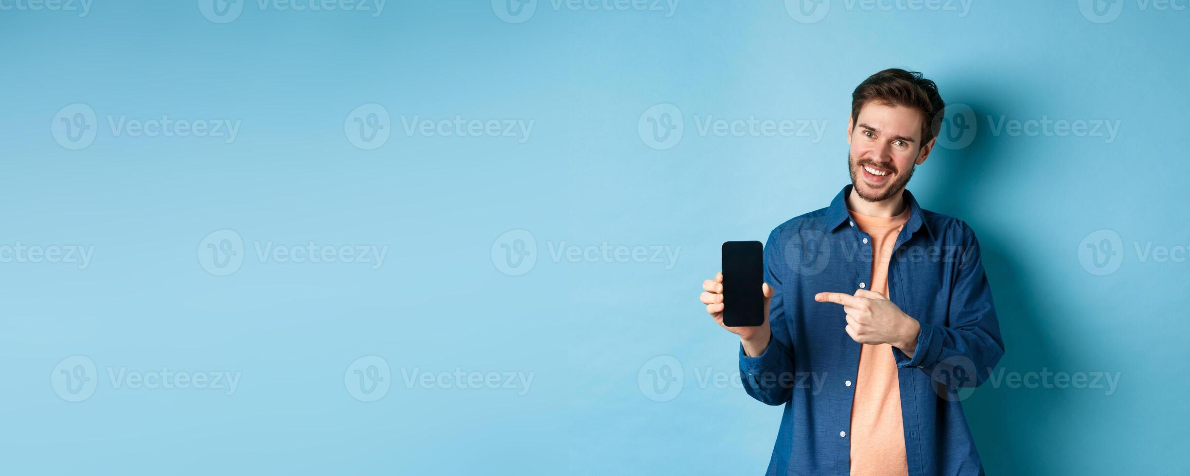 Smiling caucasian man showing on empty smartphone screen, pointing at mobile phone and looking satisfied, standing on blue background photo