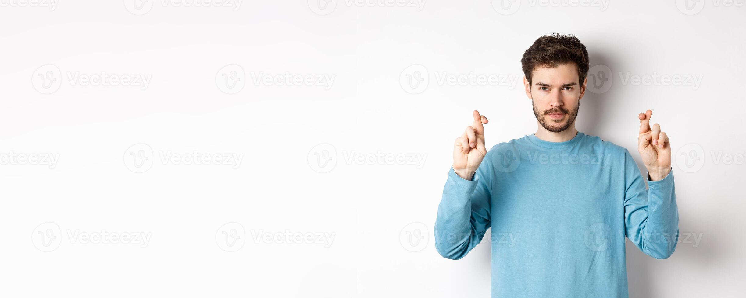 Hopeful and confident young man cross fingers for good luck, making wish or praying, anticipating results, standing over white background photo