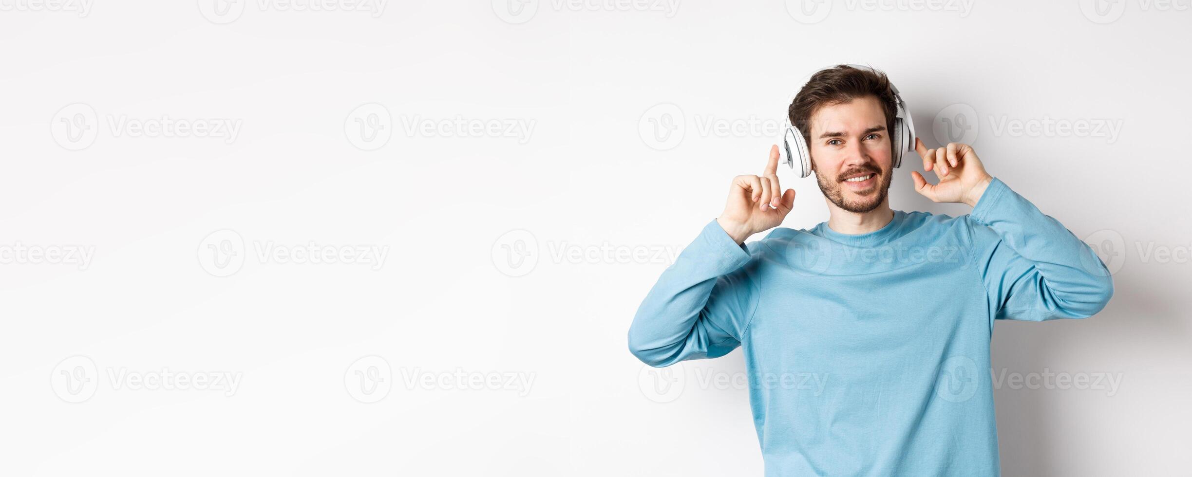 Attractive modern man with beard, smiling pleased, touching headphone on head while listening to music, standing over white background photo