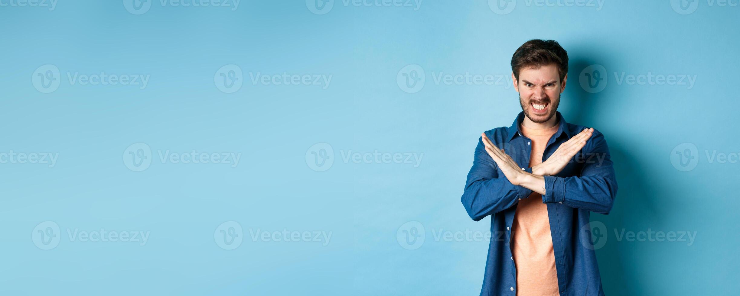 Angry young man frowning and clenching teeth outraged, showing cross gesture to stop or forbid something, standing on blue background photo